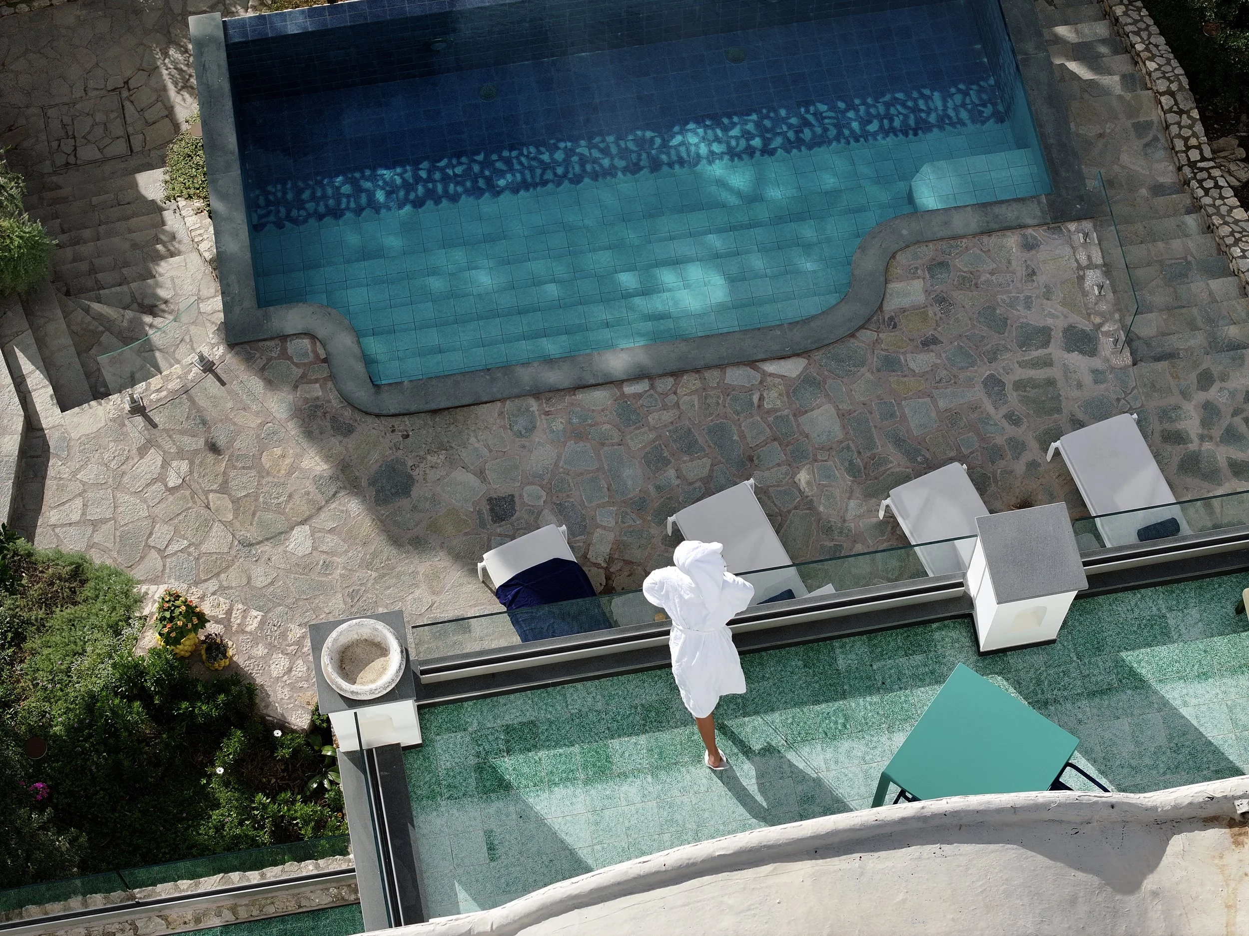 View of a swimming pool area from above, showing a woman in a white robe standing on the patio, surrounded by lounge chairs and lush greenery.