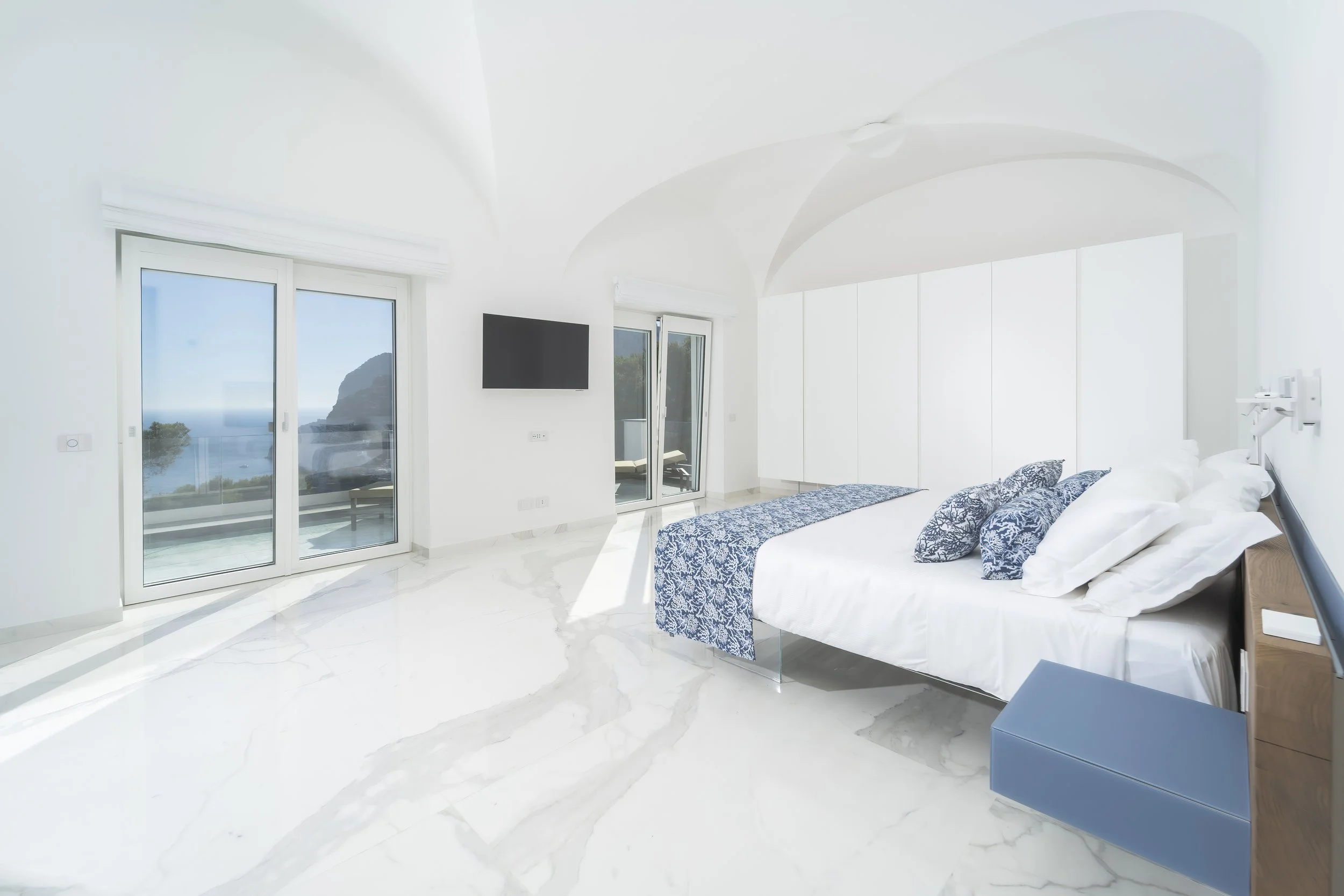 Bright bedroom with white walls and marble floor, large sliding glass doors leading to a balcony, a wall-mounted TV, a bed with white bedding and blue patterned pillows, and a scenic ocean view