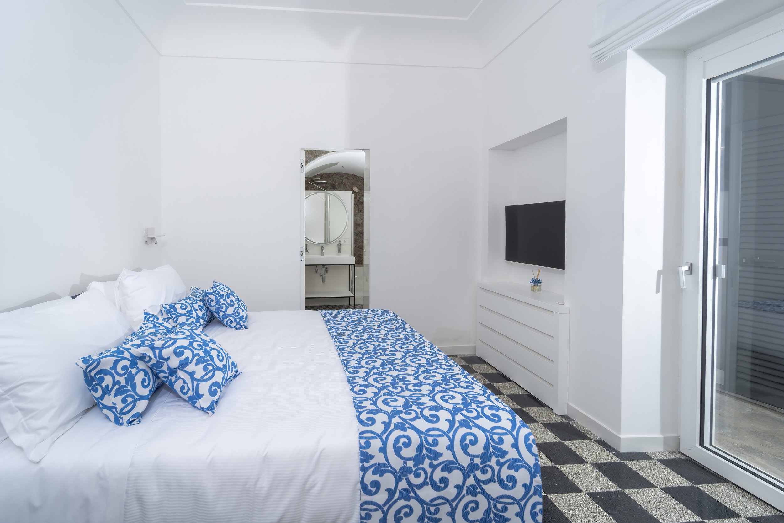 A bright white bedroom with a bed decorated with blue patterned pillows and a matching bed runner. There is a TV mounted on the wall and a sliding glass door leading outside. A bathroom with a sink and mirror is visible through an open door at the end of the room.