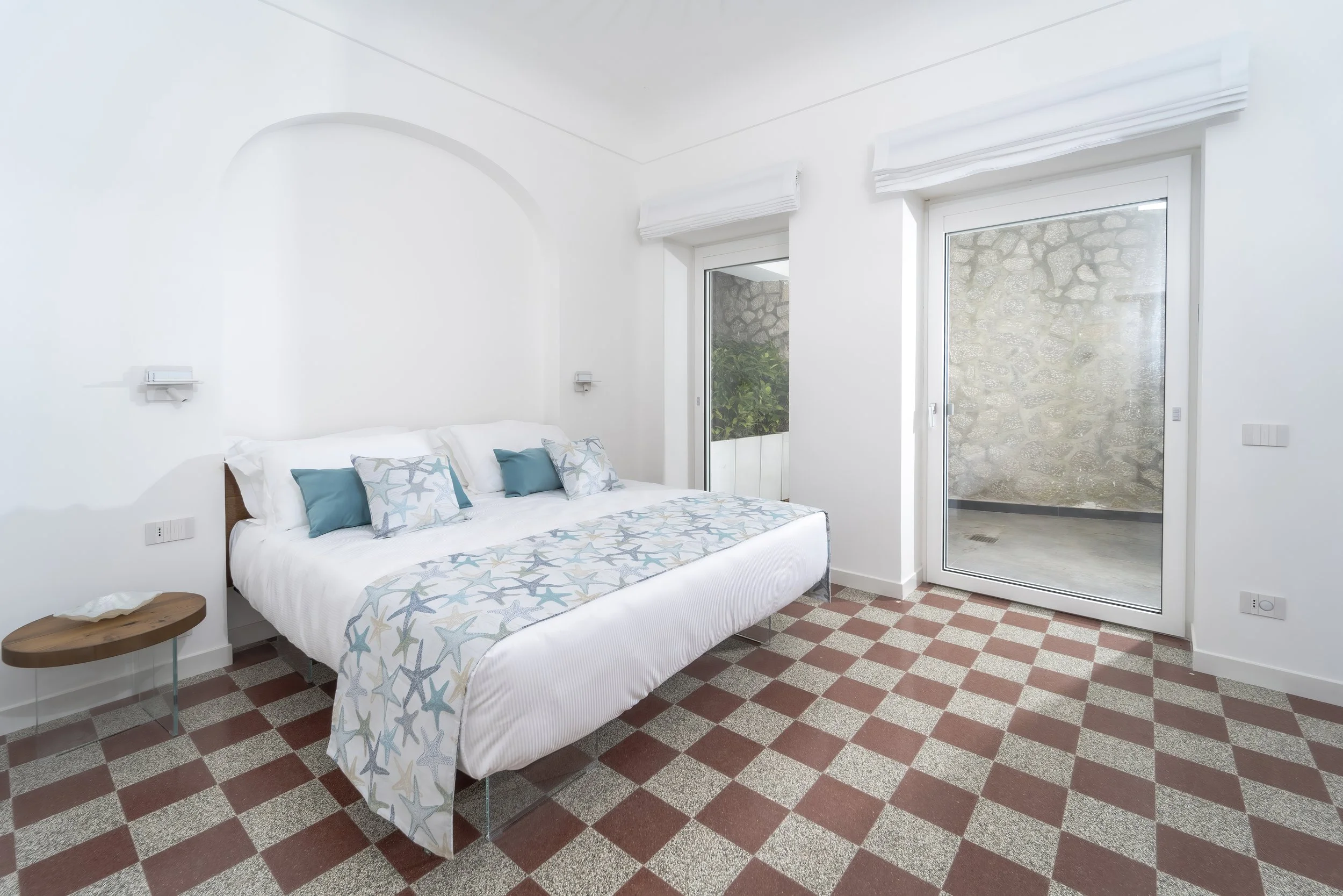 A bright bedroom with white walls, a bed with starfish-patterned bedding, teal and patterned pillows, a glass sliding door leading to an outdoor area, and checkered reddish-brown and beige tile flooring.