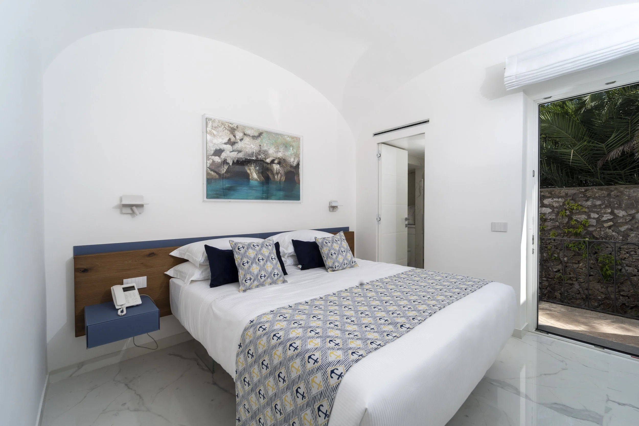 Modern bedroom with white walls, a king-size bed with a navy and white anchor patterned bedspread and decorative pillows, a blue floating nightstand with a white phone, a ceiling painting of a rocky coast with water, and a sliding glass door opening to a patio with greenery.