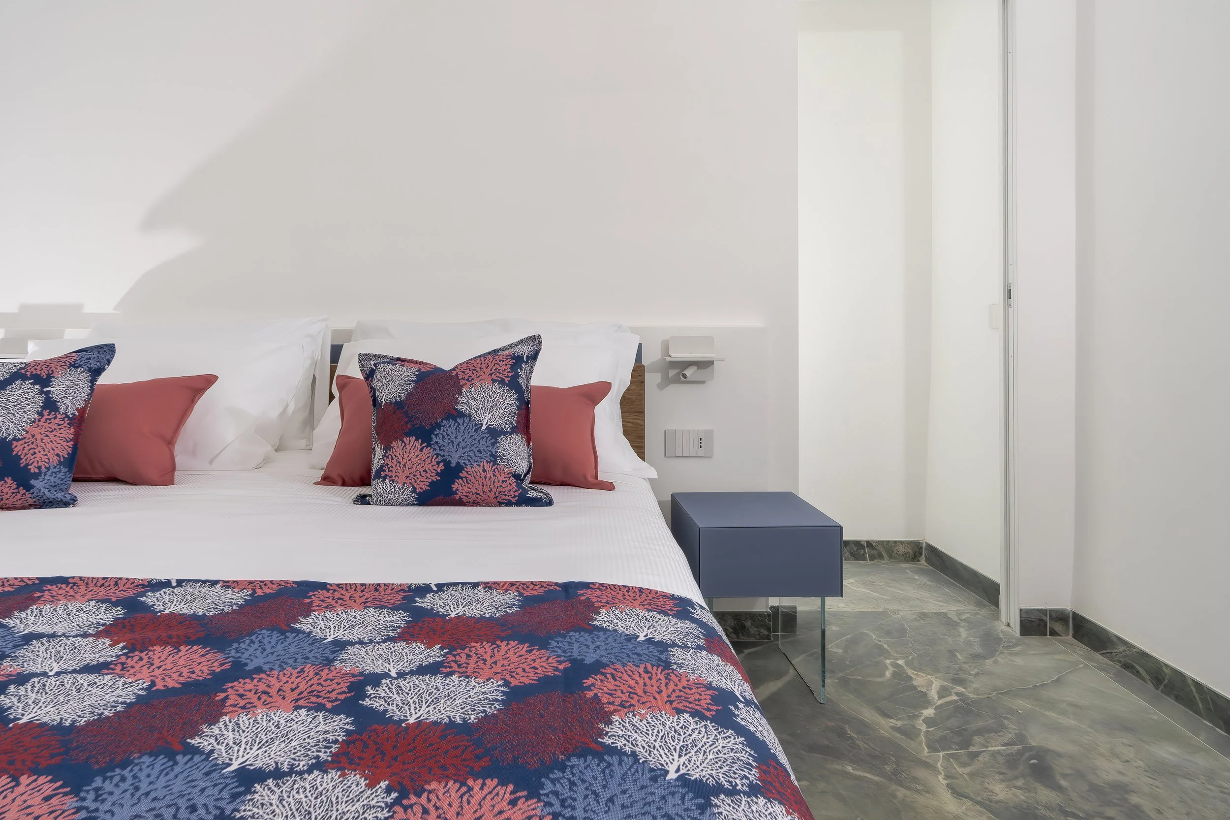 A bed with navy blue, white, and coral patterned pillows and blanket, white bedsheets, white pillows, and a small blue nightstand in a modern, minimalist bedroom with white walls and marble flooring.