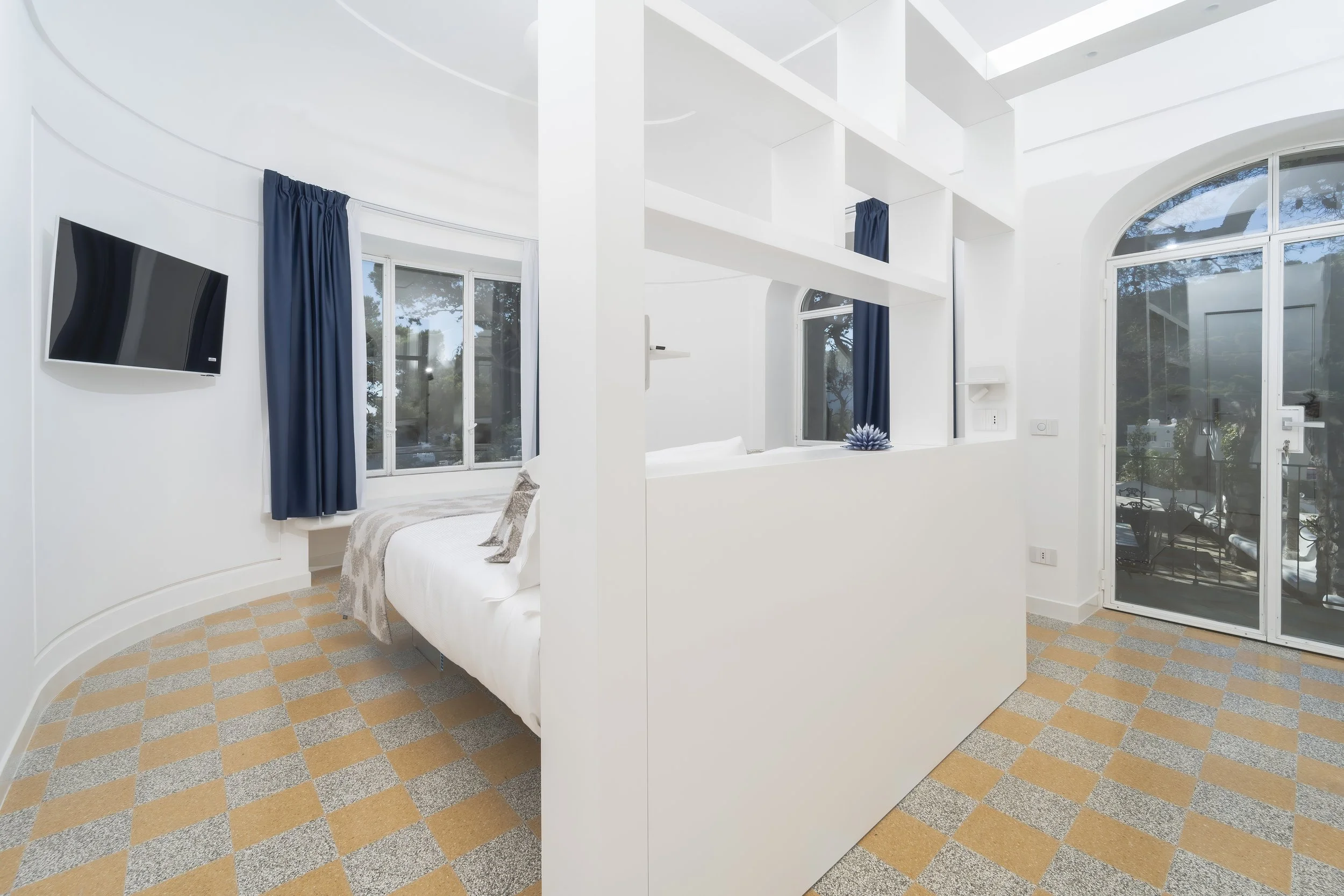 White bedroom with large windows, blue curtains, and a flat-screen TV on the wall