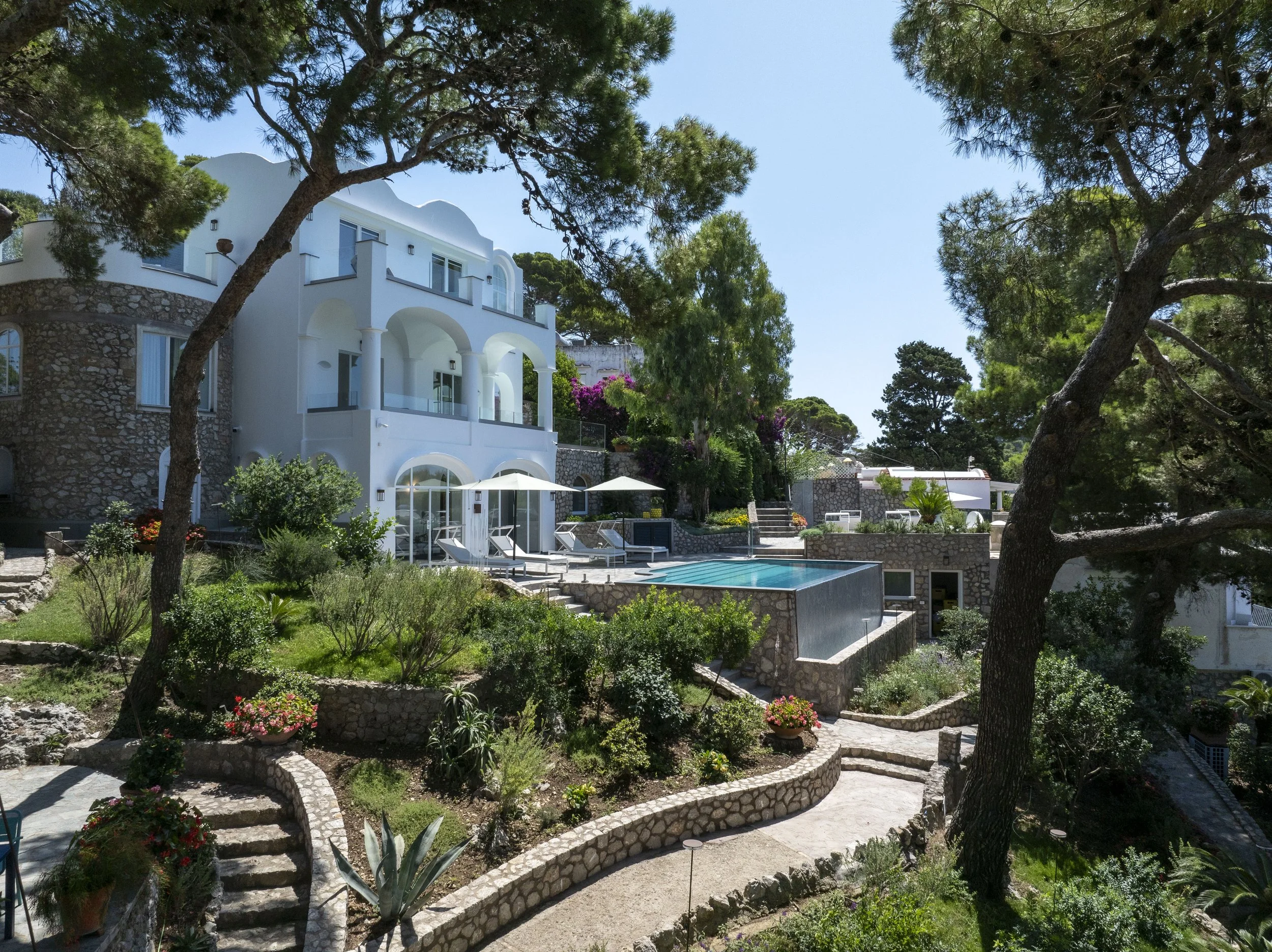 A multi-story white villa with arched windows and balconies, surrounded by lush green trees, overlooking a landscaped garden with colorful flowers, and a swimming pool with lounge chairs and umbrellas in the foreground.