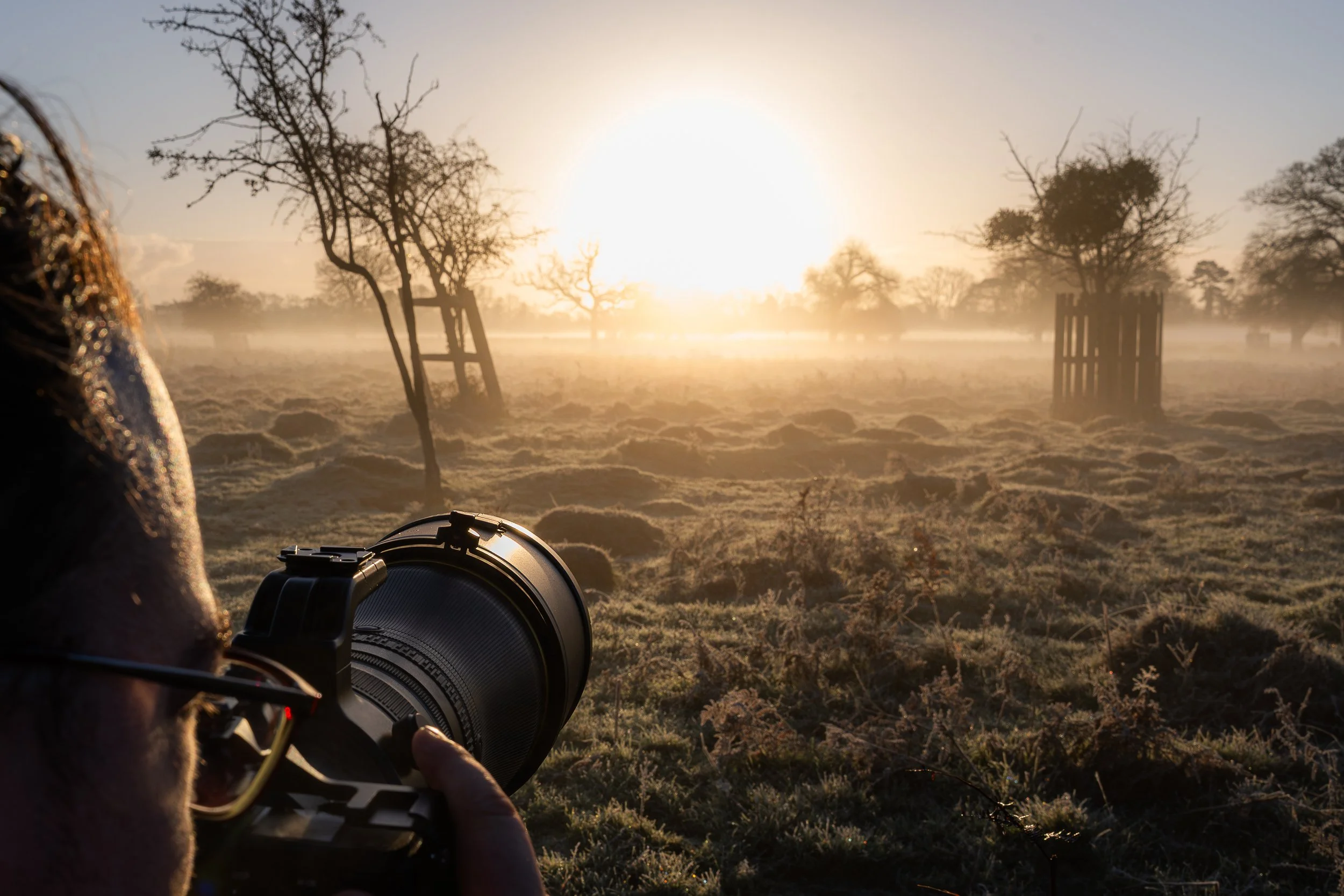 Person with glasses taking a photograph of a sunrise over a frosty field with sparse trees and wooden structures.