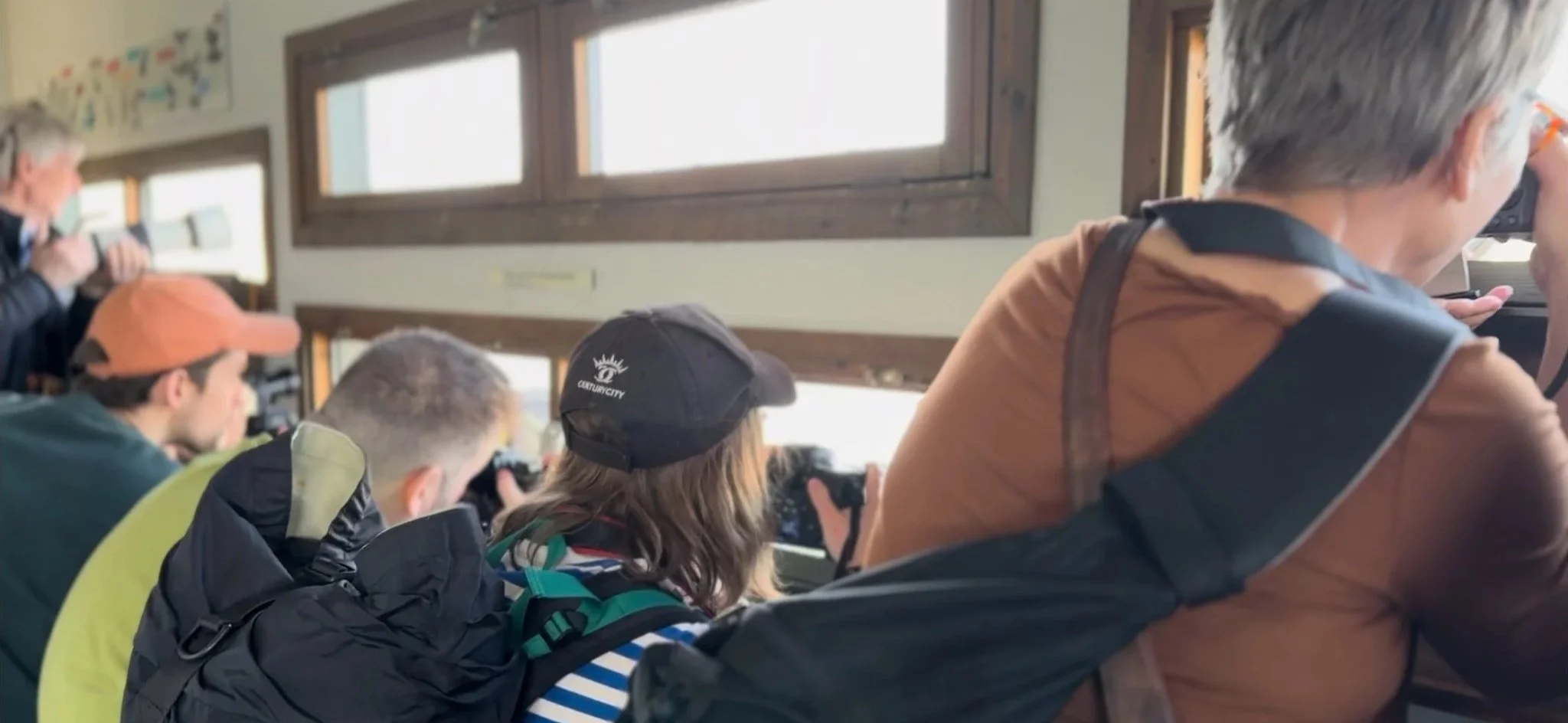 Several students in a classroom observing through binoculars and taking photos with cameras, sitting near windows.