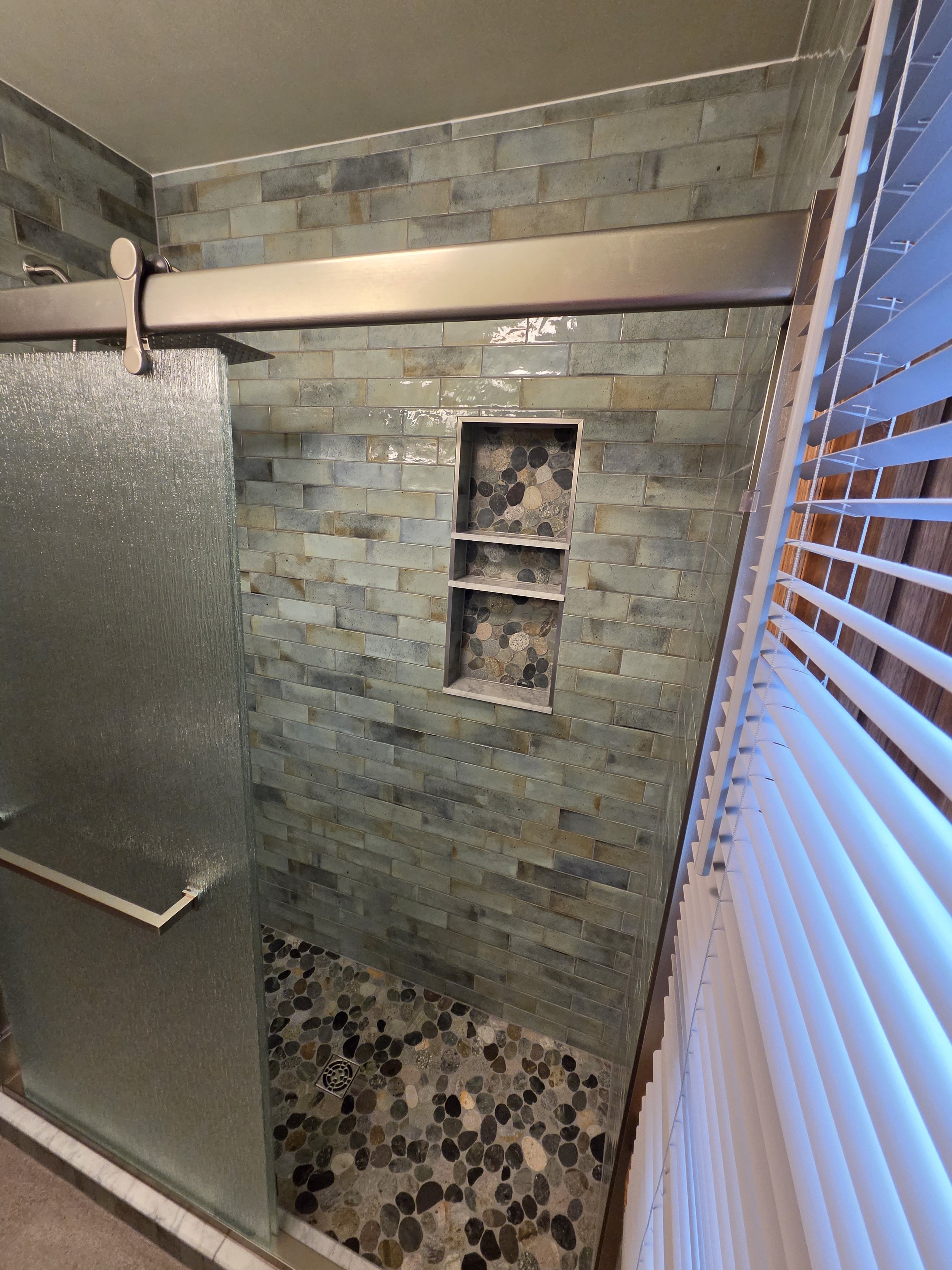A modern shower with green subway tiles, a pebble stone floor, a glass door, a recessed shelf with pebbles, and a window with white blinds.