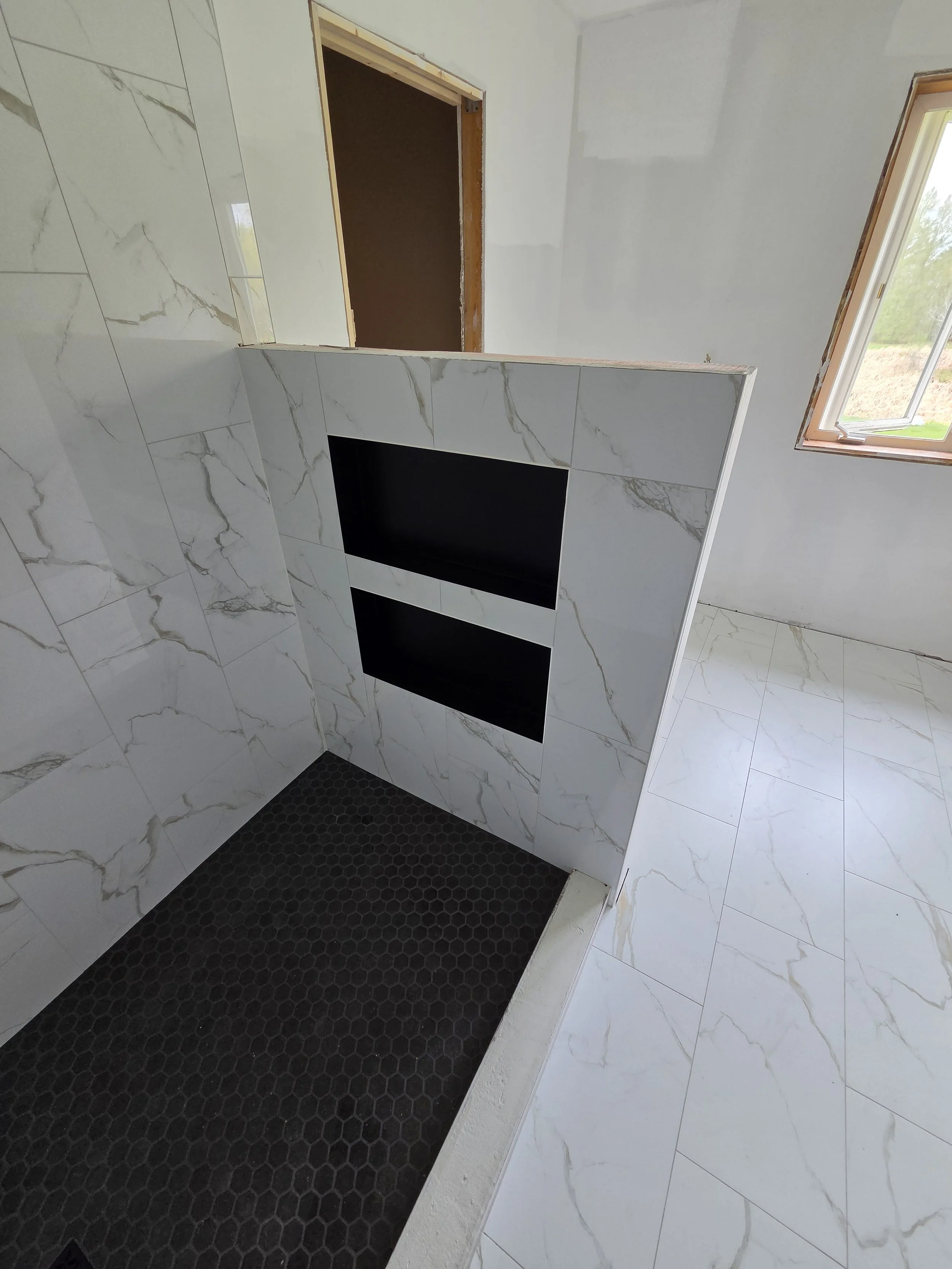 Bathroom under construction with marble tiles on the walls and floor, a black shower area with hexagonal tiles, a window, and an opening for a door.