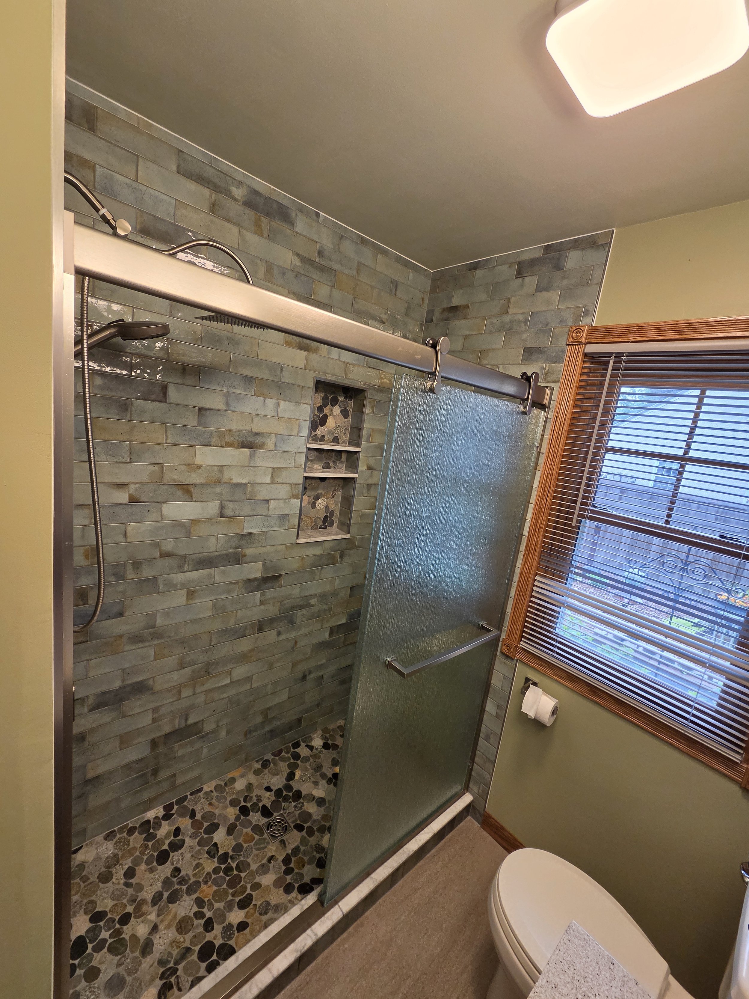 Bathroom with a green tiled shower area and a sliding frosted glass door. The shower has built-in shelves with pebble stone decor and a handheld showerhead. A window with wooden blinds is on the right, and a toilet is partially visible at the bottom 