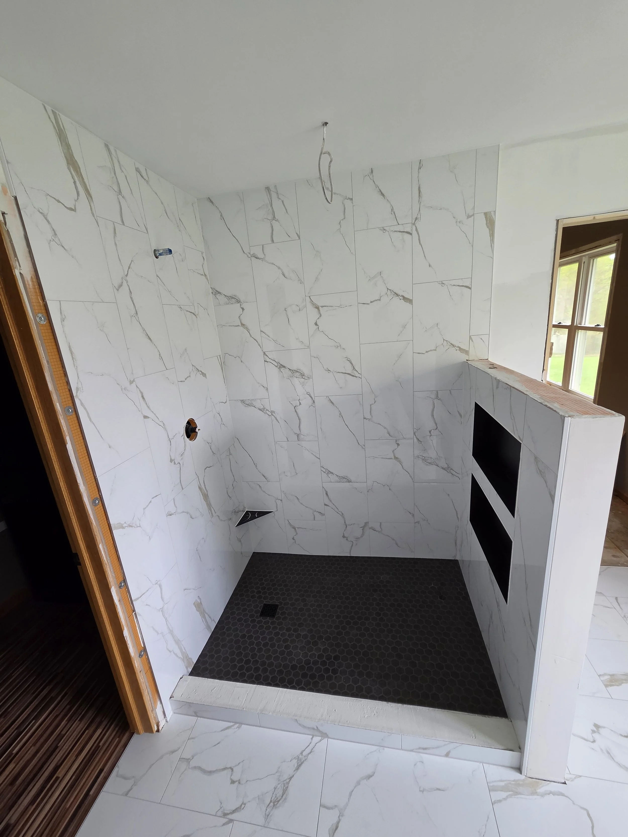 View of a bathroom shower area with white marble tiles, black hexagonal floor tiles, and a small window.
