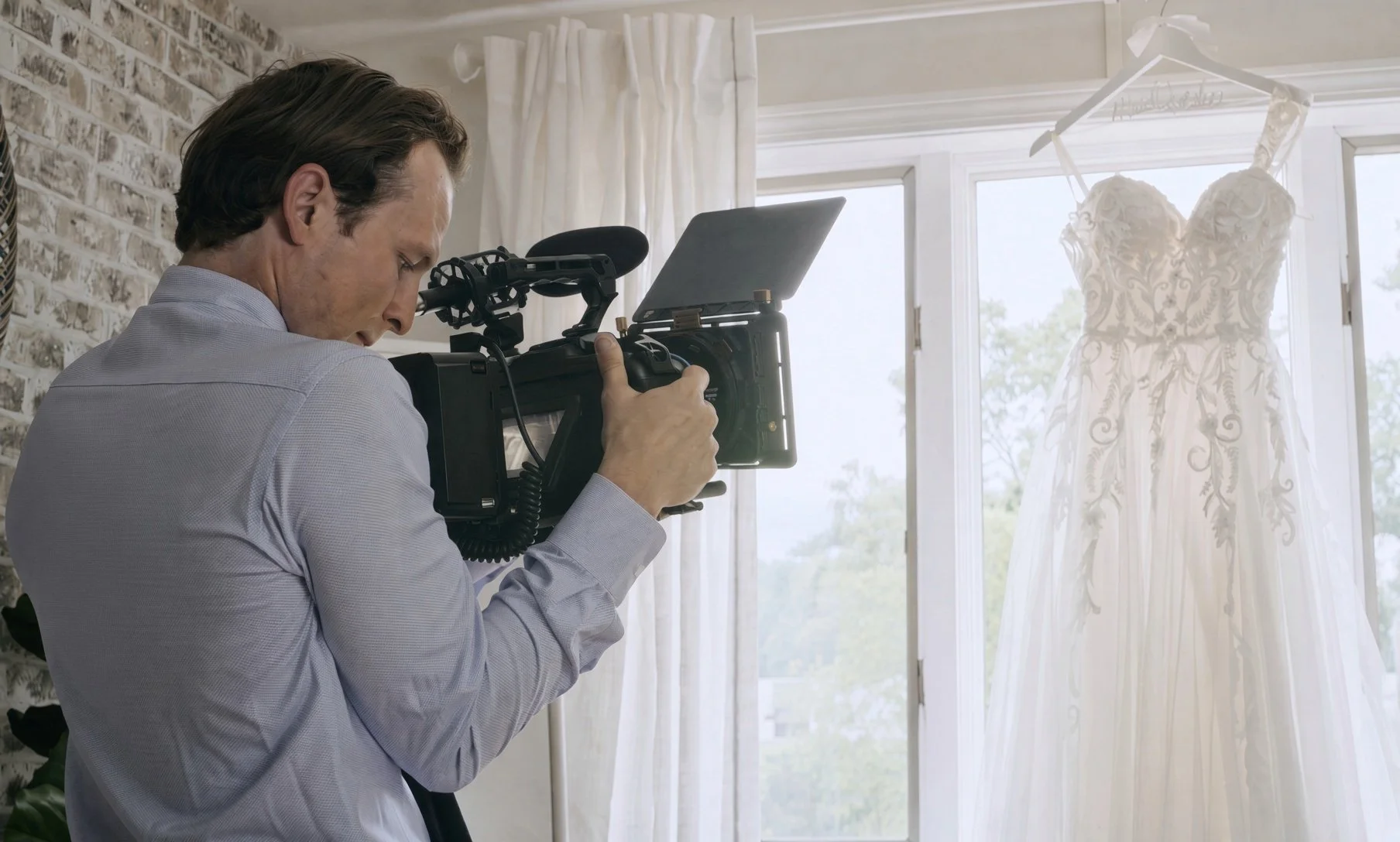 Videographer filming an ornate white wedding dress hanging by a window.