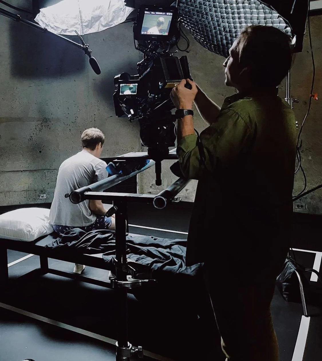 A film crew filming a scene with a young man sitting on a bed in a dimly lit room, using professional lighting and camera equipment.