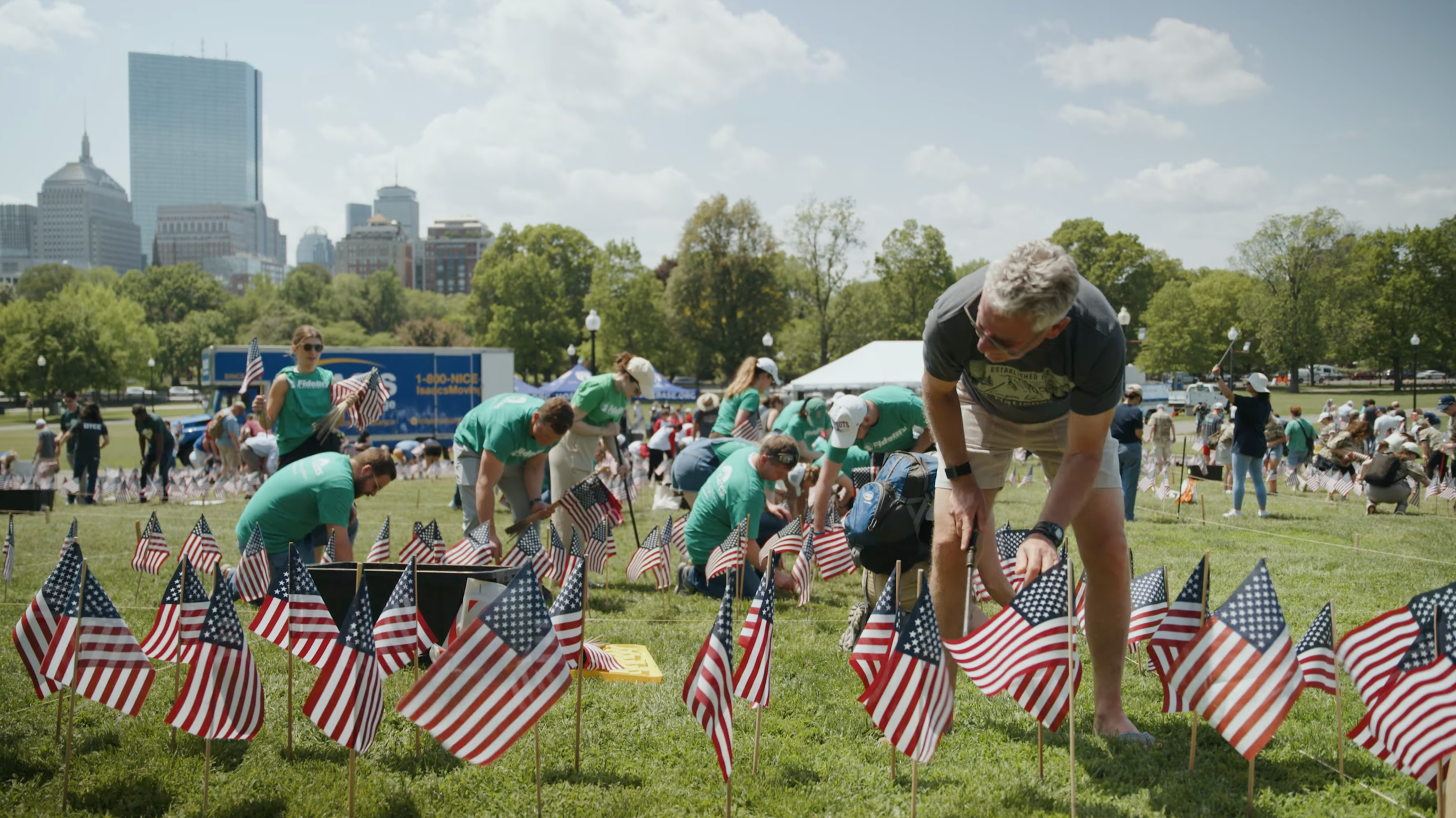 Volunteers plant rows of American flags on Boston Common for Home Base’s 2026 Memorial Day Flag Garden, with the Boston skyline behind them.