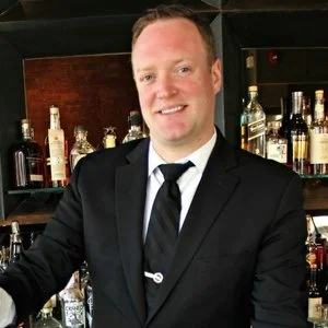 Man in a suit standing in front of a bar with various bottles of liquor.