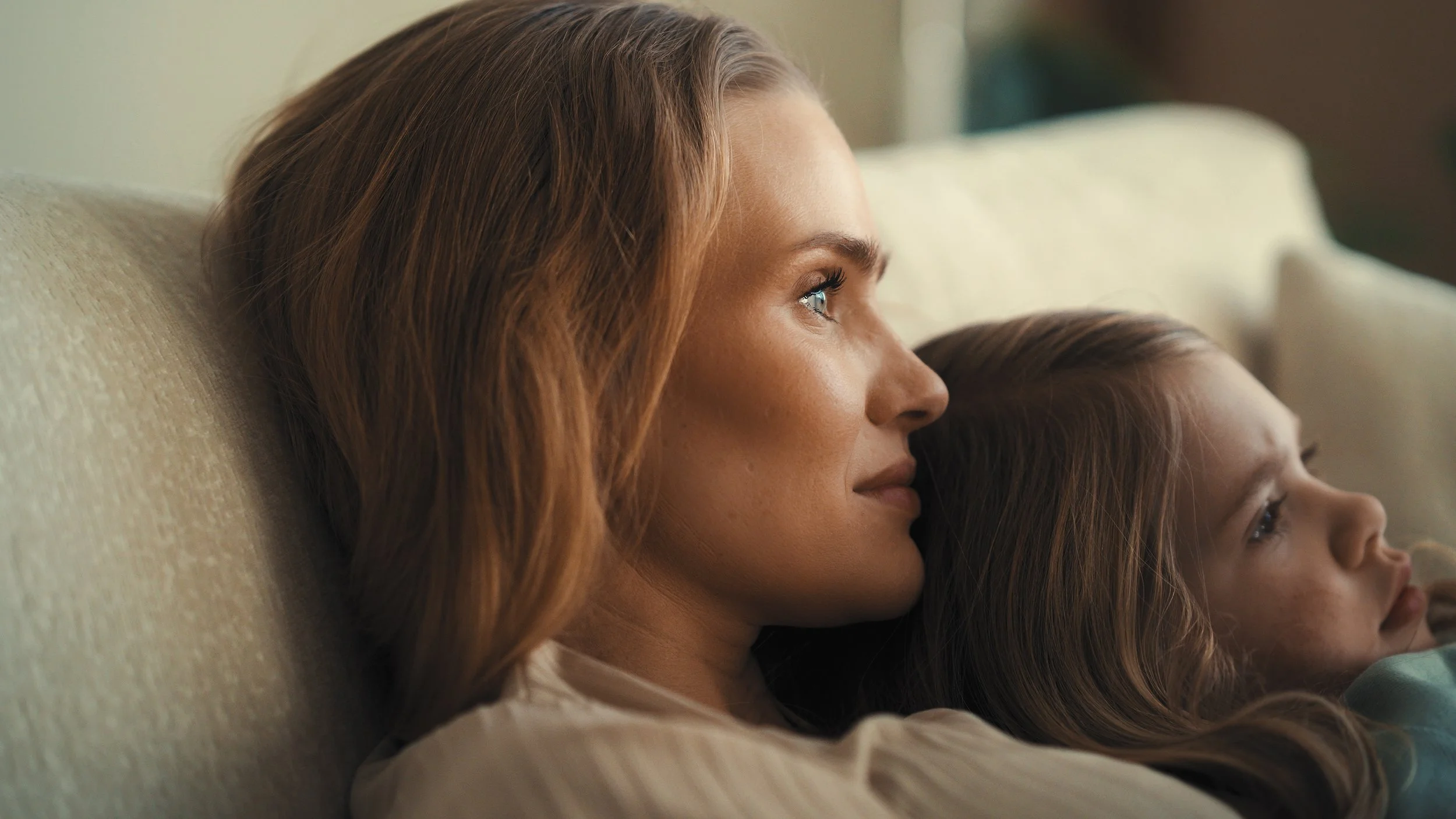 A woman with reddish hair and a young girl with brown hair, both lying on a beige couch, looking peacefully at something off-camera.