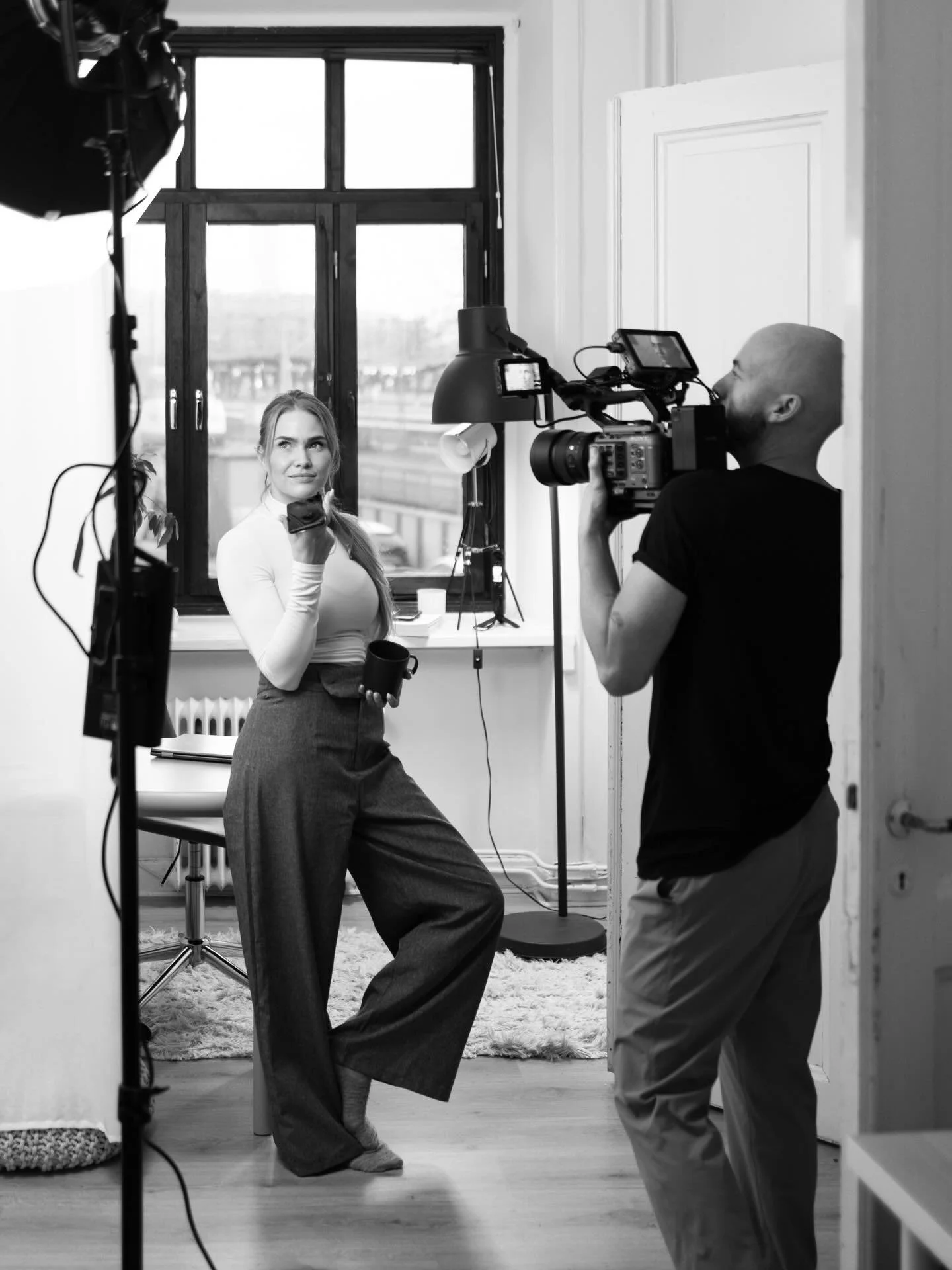 A woman in a white top and high-waisted pants holding a coffee mug and makeup brush, being filmed by a cameraman in a room with a large window and a soft rug.