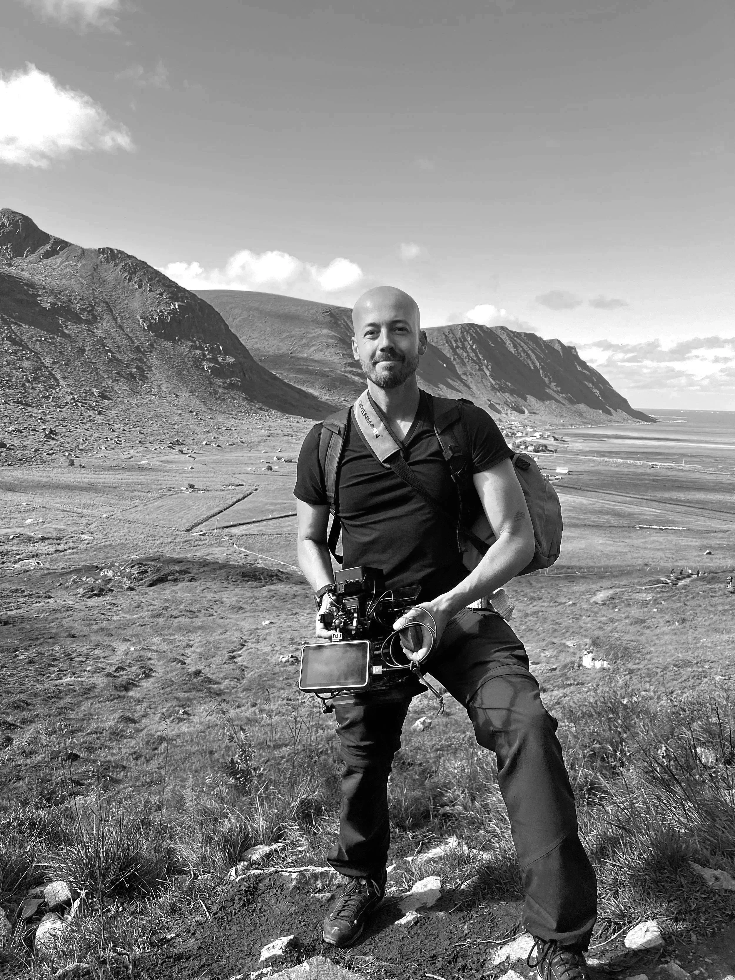 A man with a backpack and camera kneeling on rocky ground in a mountainous landscape with cliffs and fields, black and white photo.
