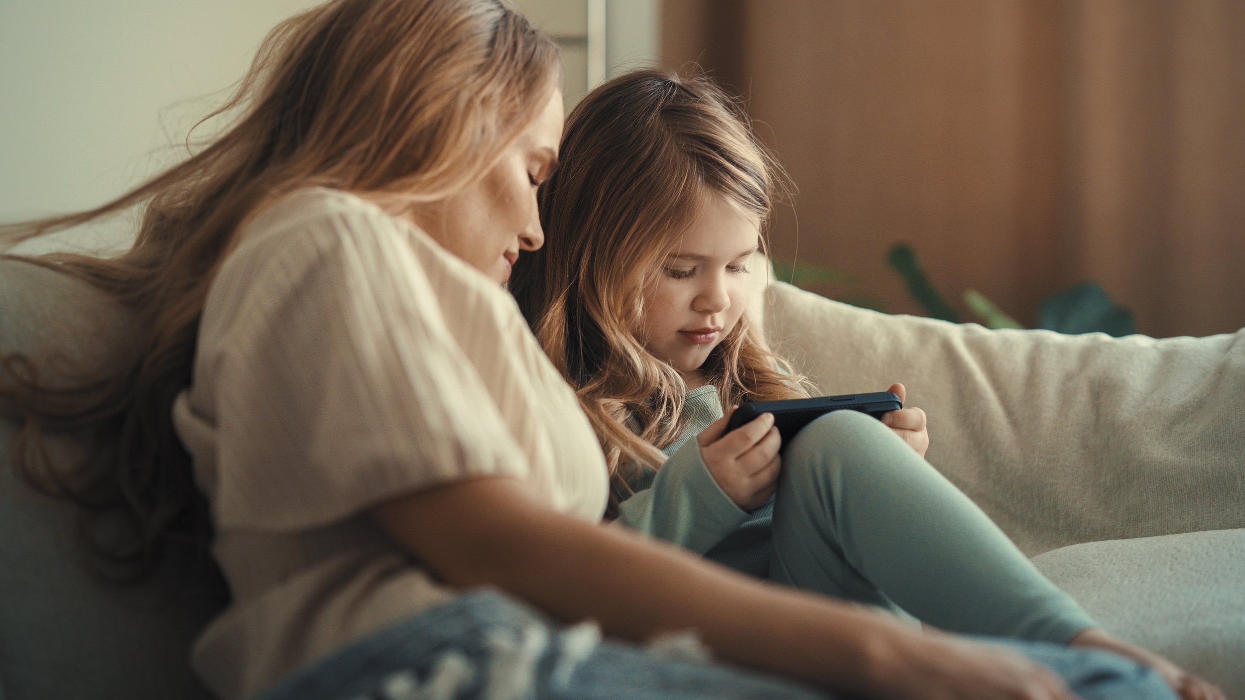 A woman and a young girl sitting on a beige couch, looking at a handheld gaming device together, in a cozy living room.