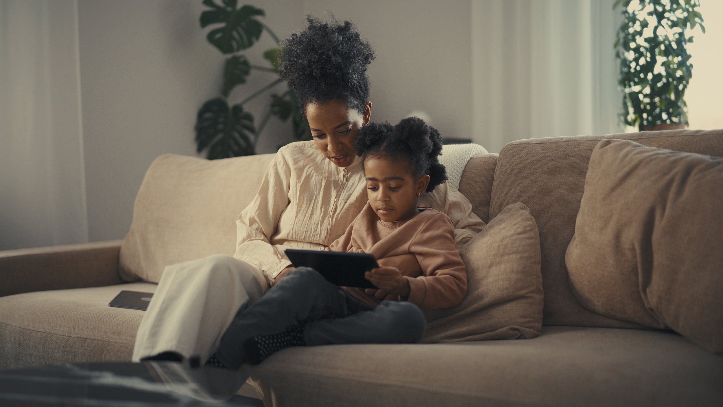 A woman and young girl sitting on a beige sofa, looking at a tablet together in a cozy living room with plants in the background.