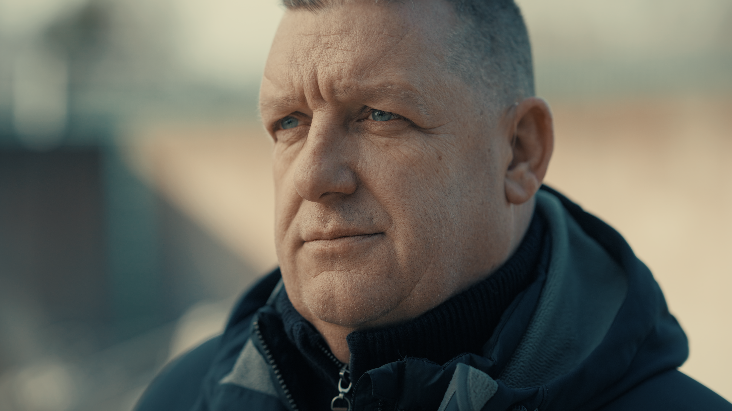 Close-up of a middle-aged man with short hair and blue eyes, wearing a black jacket with a zipper and a dark blue shirt underneath. He is outdoors with a blurred background.