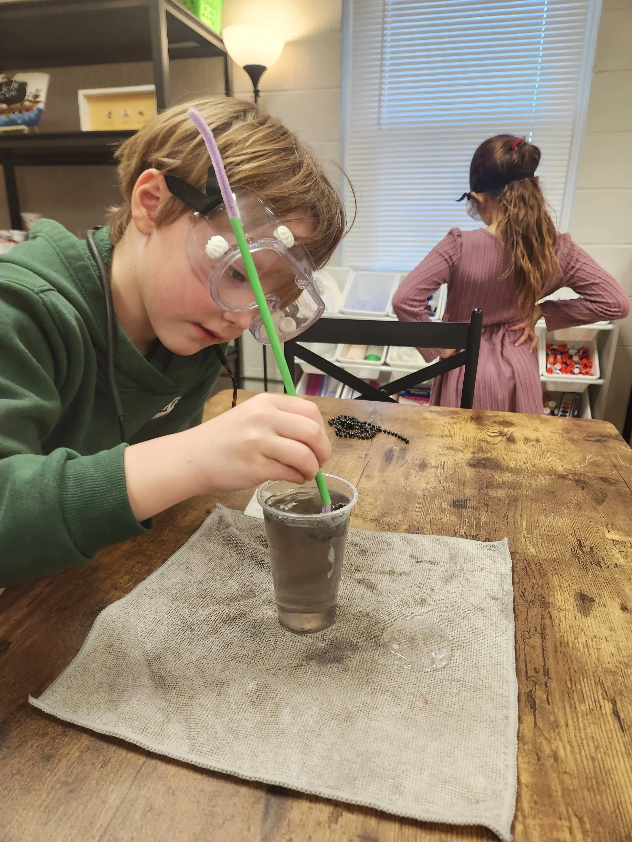 A boy with goggles and a green hoodie is placing black beads into a clear cup of water with a green straw. A girl with a red tie and pink dress stands in the background near a storage shelf. They are completing a hands-on science experiment.