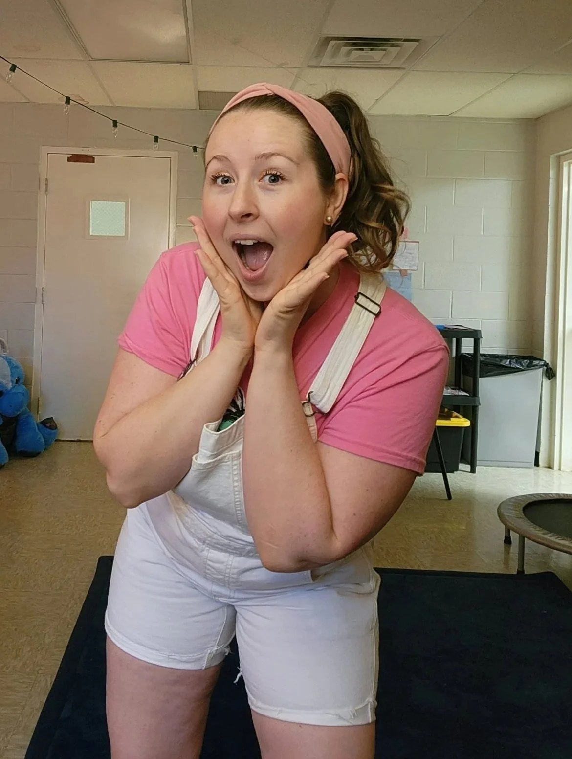 A woman with a pink headband, pink t-shirt, and white overalls posing indoors with surprised and happy expression. She is the founder of Change Makers Academy, a microschool in Nashville, Tennessee.