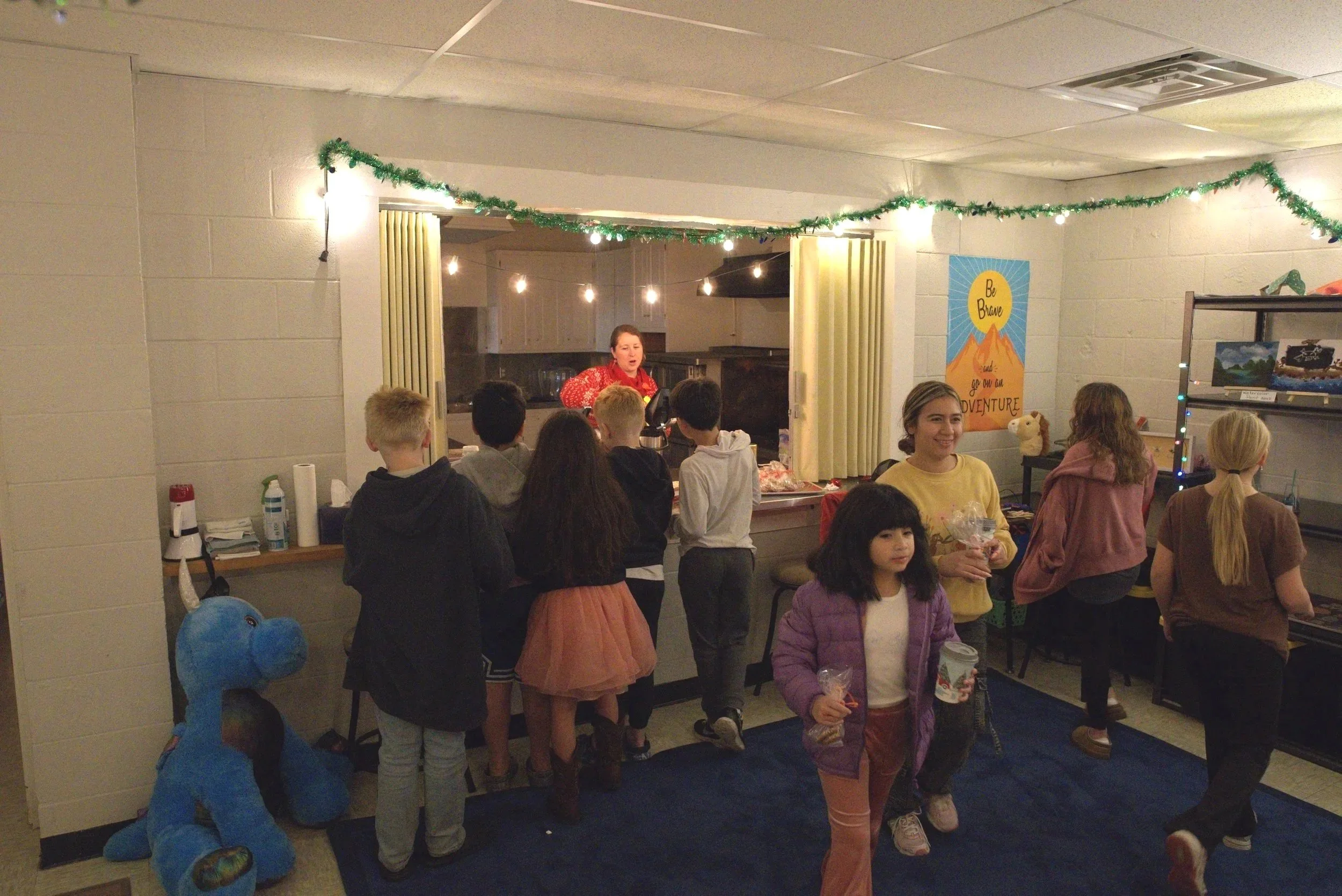 Children gather around a kitchen area at Change Makers Academy, with a woman in red in the background who is serving hot cocoa. Decorations include green garland and a poster that says 'Be Brave, Go on an Adventure'.