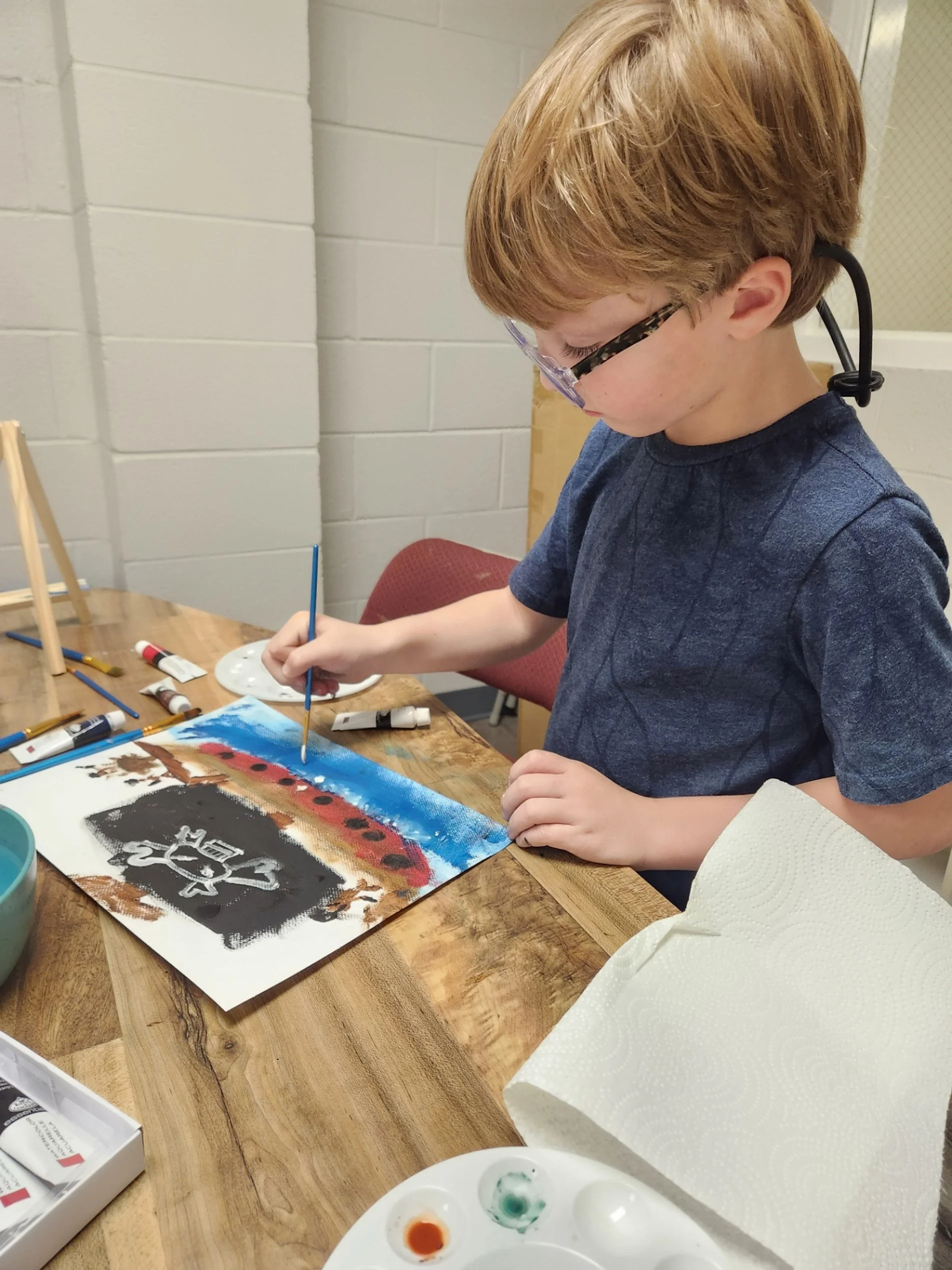 A young boy with blonde hair and glasses painting a colorful artwork of a pirate's ship on paper using watercolors, sitting at a wooden table. The location is a microschool in Tennessee.
