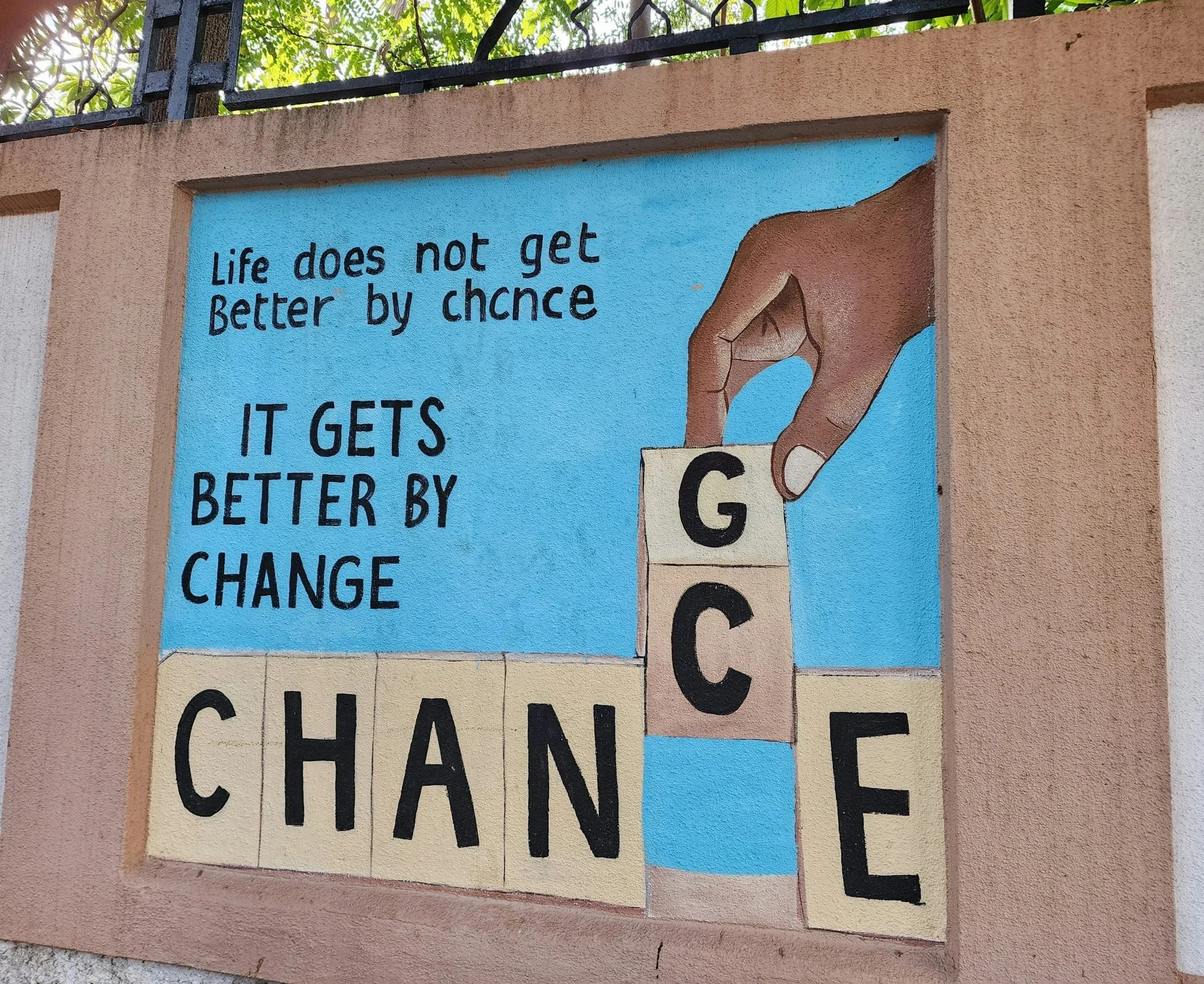 A painted mural on a wall featuring a hand placing letter tiles to spell the word 'CHANGE'. The mural includes a quote: 'Life does not get better by chance, it gets better by change.' The background of the mural is blue, and the wall is pinkish with a black metal fence above it.