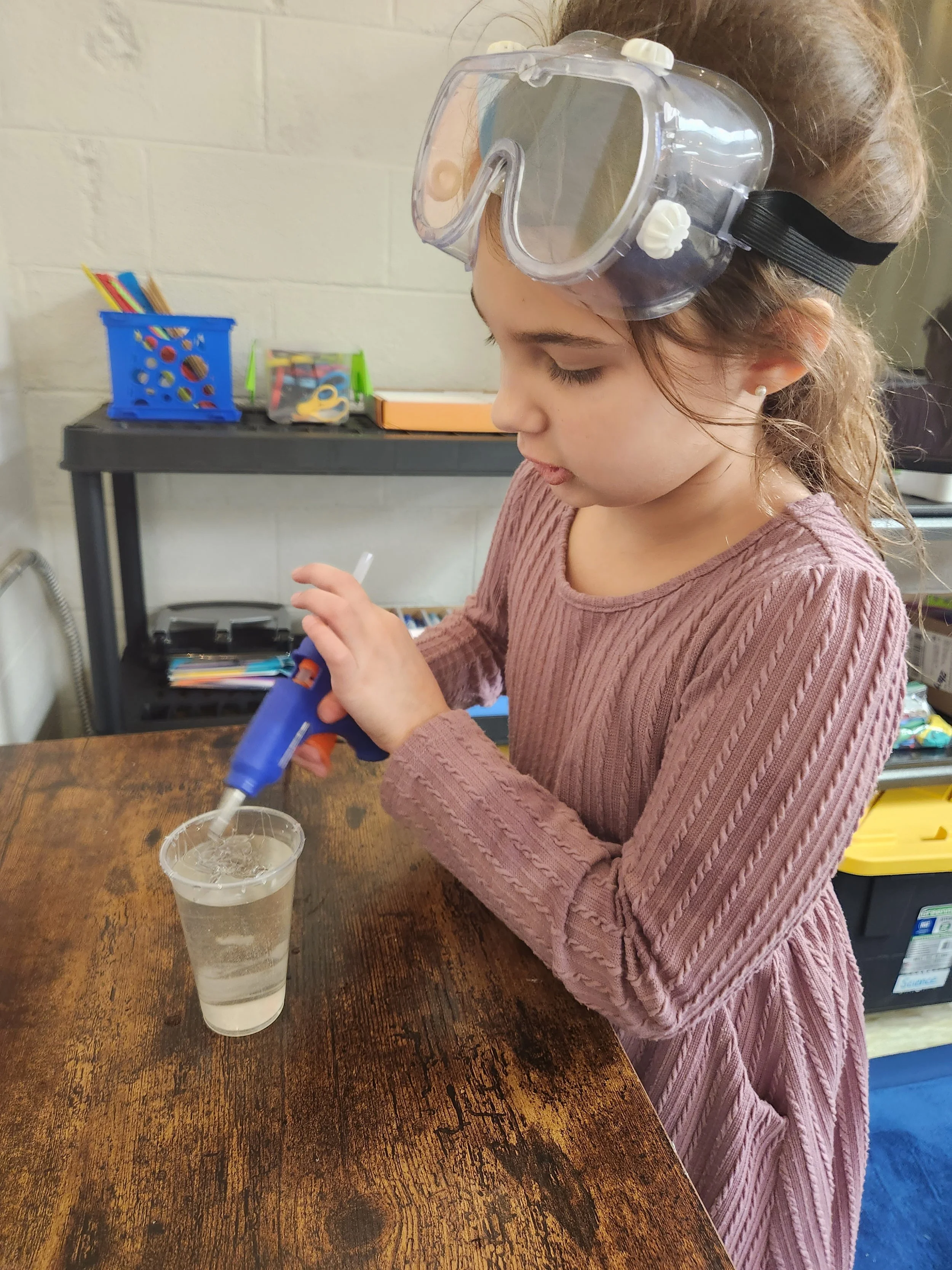 Young girl in pink sweater wearing safety goggles, using a hot glue gun to apply glue to a clear plastic cup on a wooden table at a microschool in Nashville.