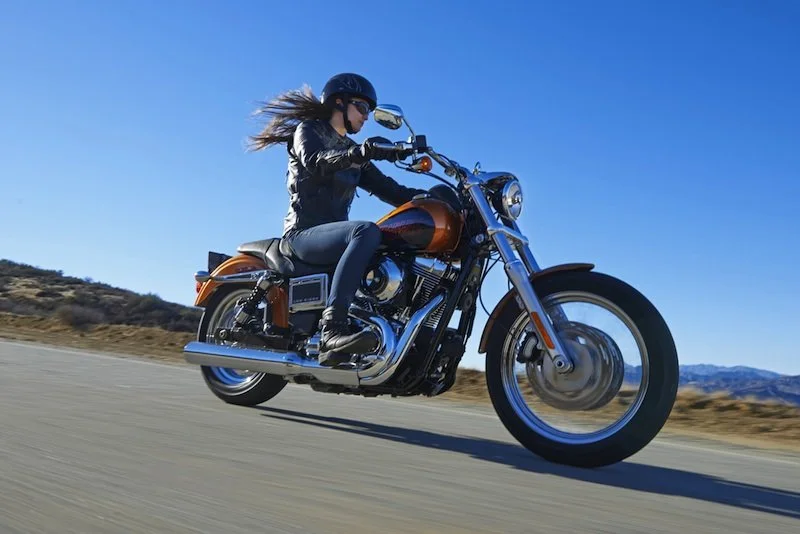 Person riding a motorcycle on an open road in a scenic landscape, wearing a helmet and leather jacket.