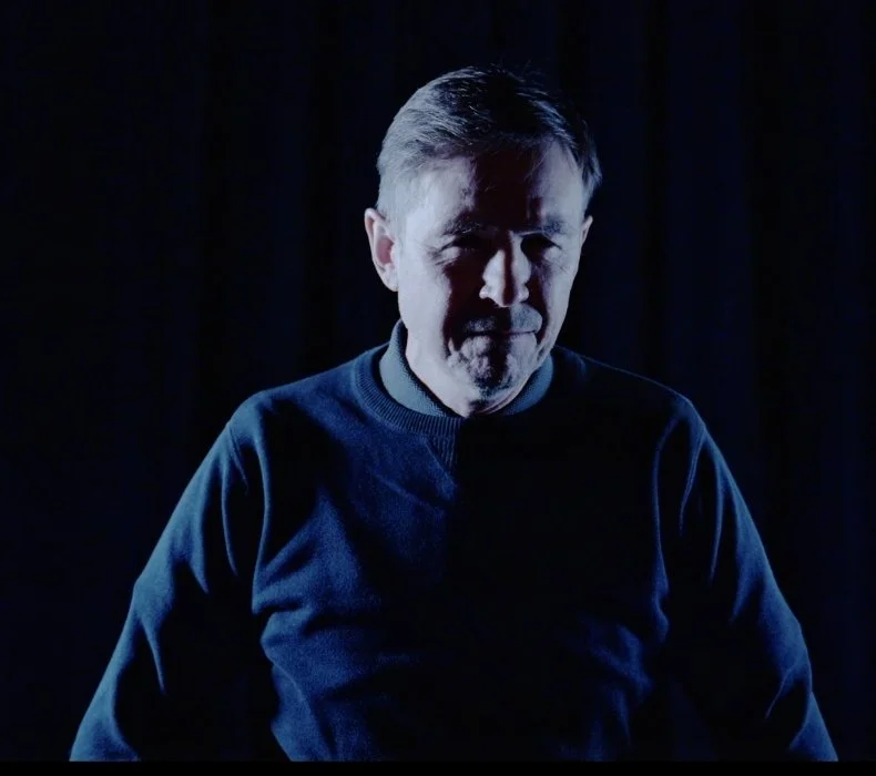 A man with gray hair and a gray beard, wearing a dark blue sweater, stands against a dark background. His face is partially shadowed, giving a mysterious or serious expression.