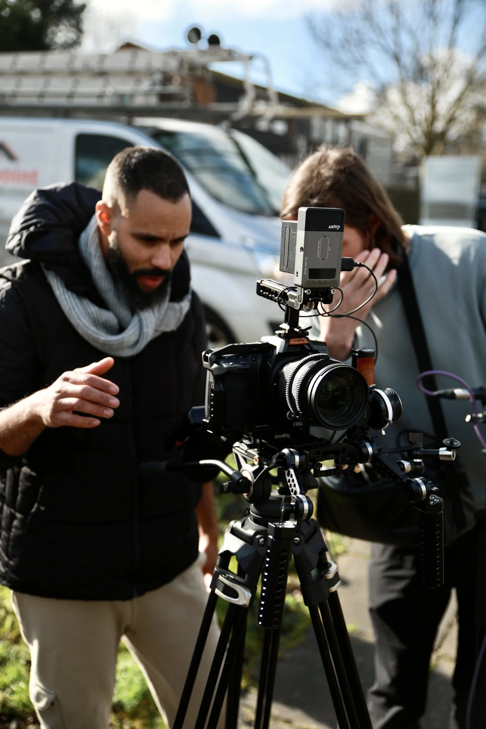 Two people are outdoors, closely inspecting a professional camera mounted on a tripod. One person, with short dark hair and a beard, is wearing a black jacket and a white scarf. The other person, with long brown hair, is wearing a light gray jacket. A truck with roofing ladders on top is visible in the background, along with a tree and a residential neighborhood.
