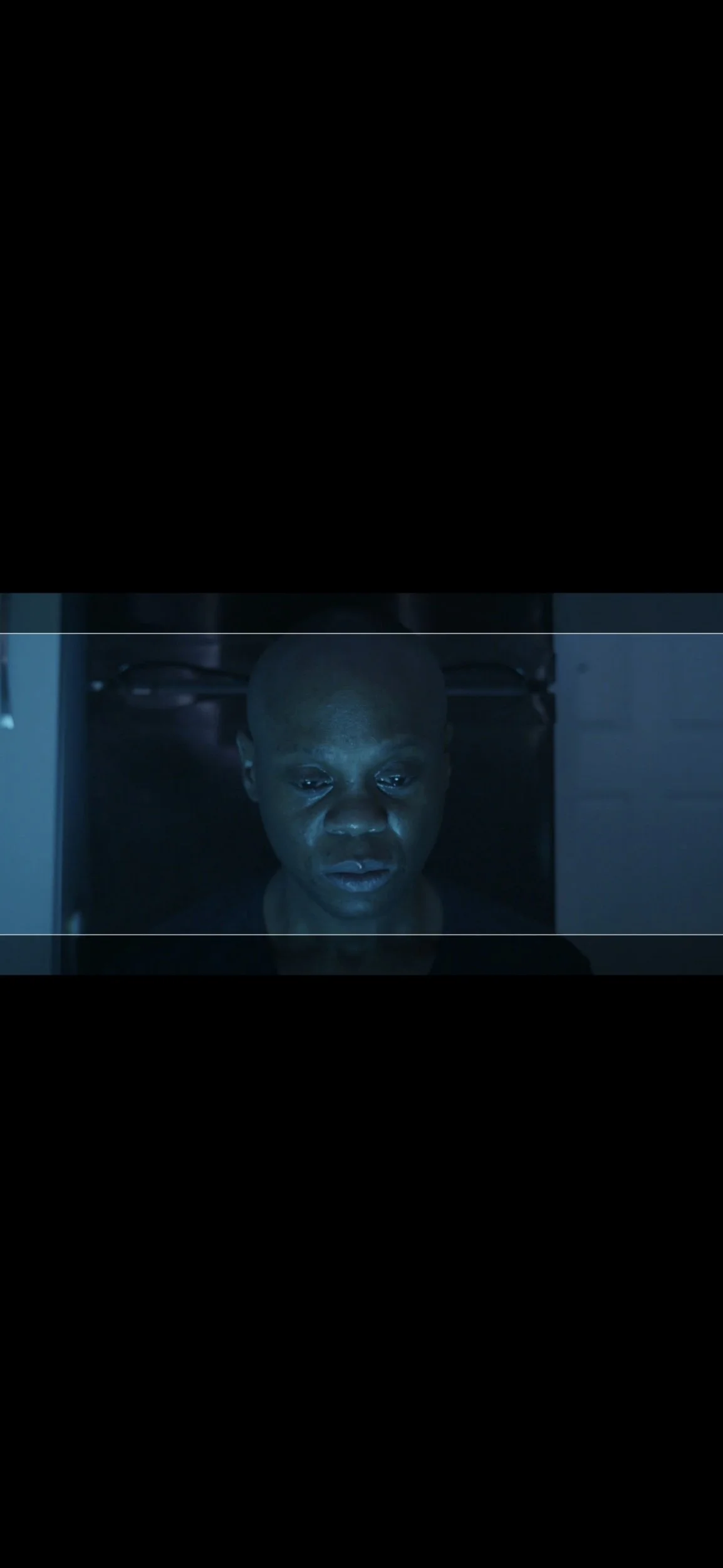 A person with a shaved head looking at a computer screen with a serious expression, illuminated by a bluish light.