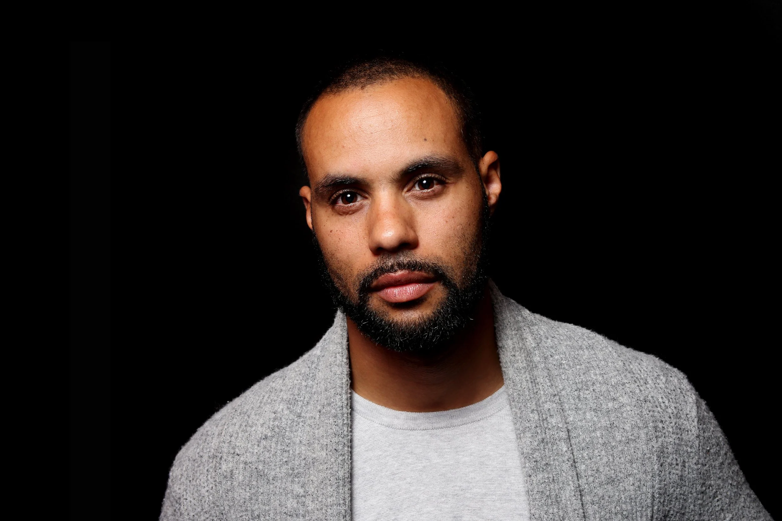 Wayne Nelson's headshot. portrait of a man with dark hair and beard wearing a grey sweater against a black background.