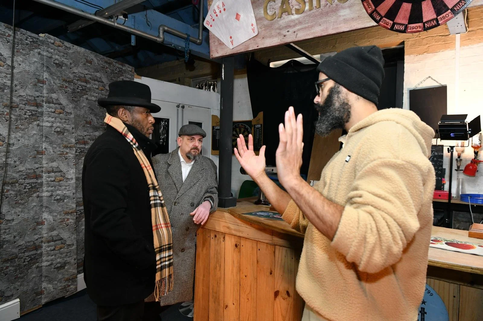 On a film set. Three men engaged in conversation inside a bar, with one man gesturing with his hands, one wearing a black hat and a scarf, another in a plaid coat, and the third in a hoodie and glasses, with a dartboard on the wall behind them.