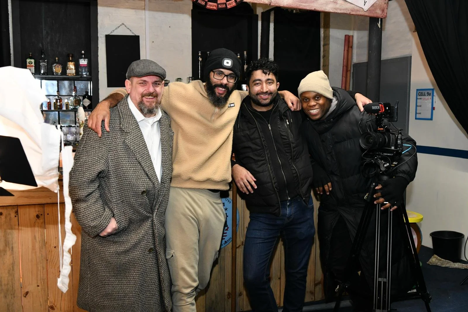 On set. Four men standing together indoors, smiling, with a camera on a tripod in front of them. Background shows bottles on shelves and a black curtain.