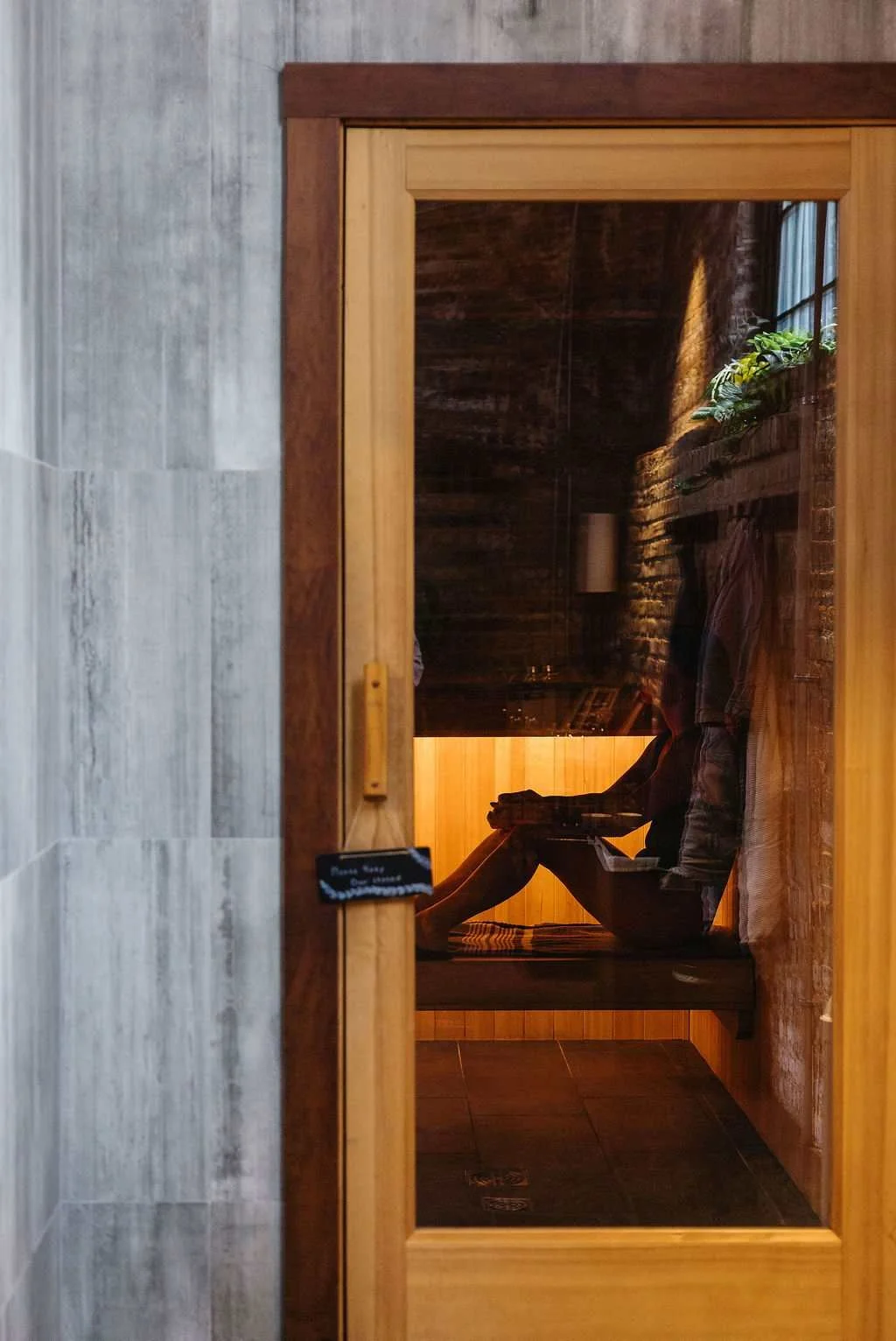 Person sitting inside a wooden sauna or steam room, reflected in a glass door, with plants and a brick wall visible inside.