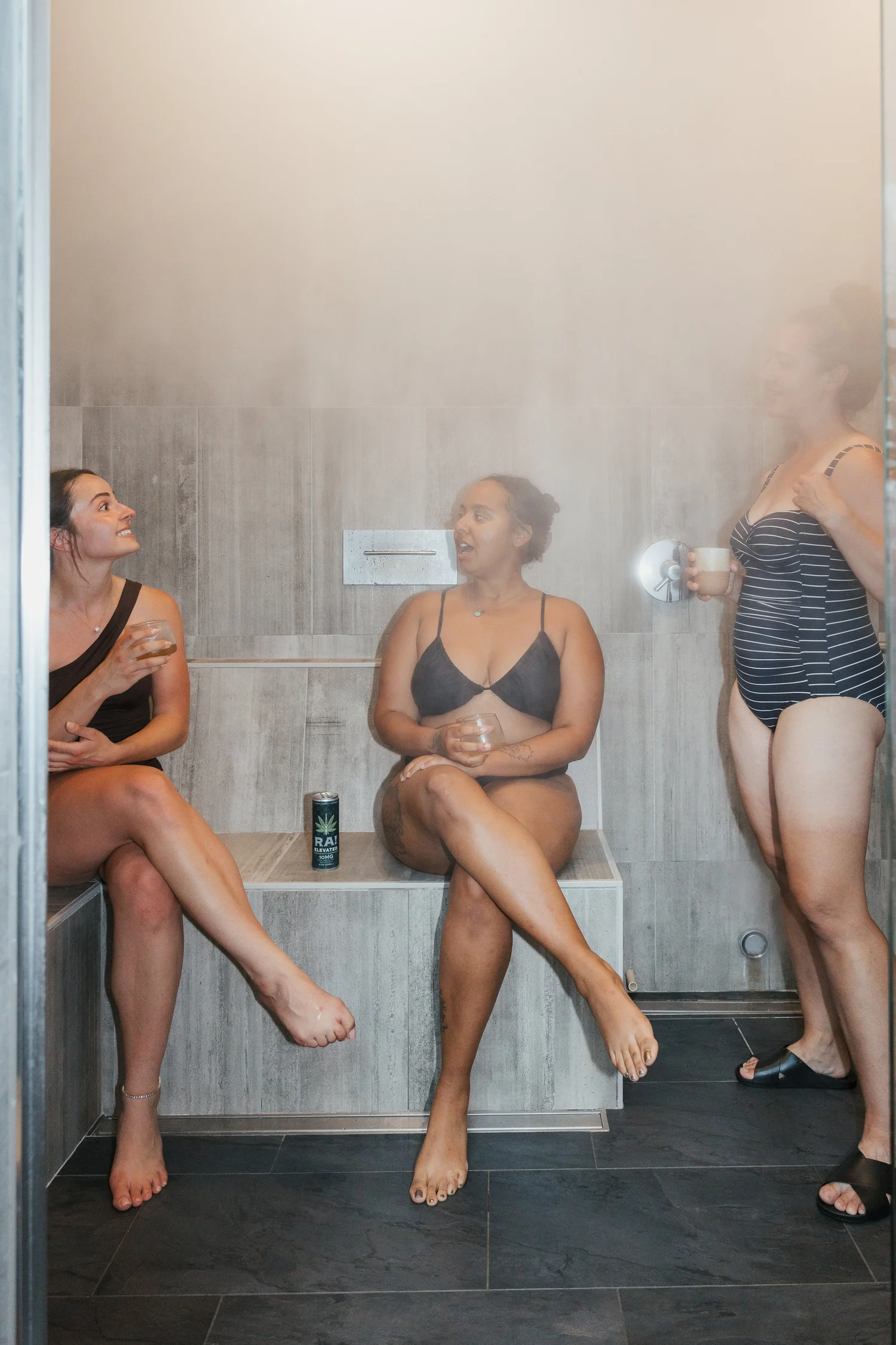 Three women relaxing in a steam room, sitting on a bench, engaged in conversation, holding cups, with a can of cannabis beverage on the bench.