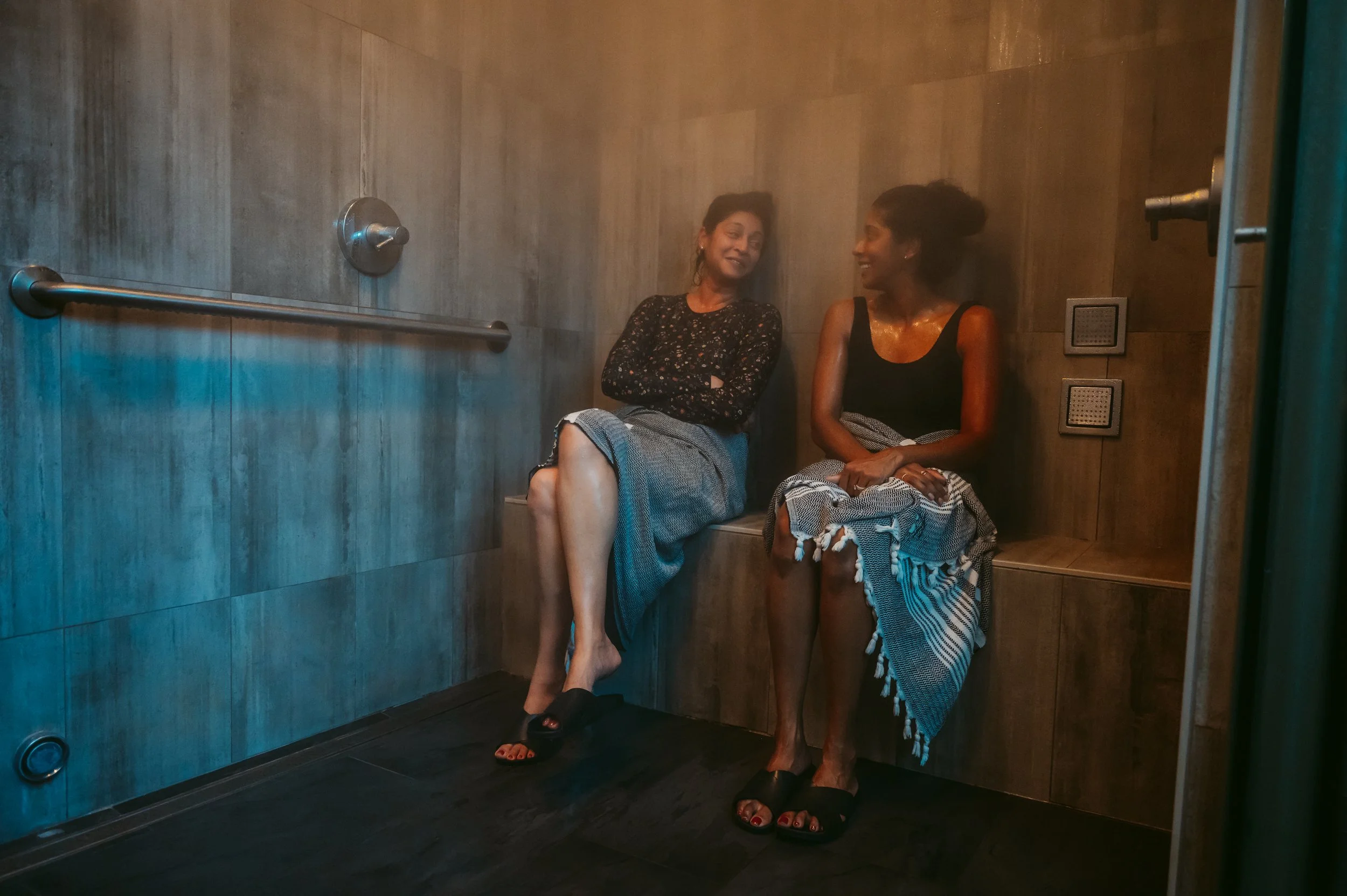 Two women sit on a bench in a steam room, wearing bath towels, and are smiling at each other.