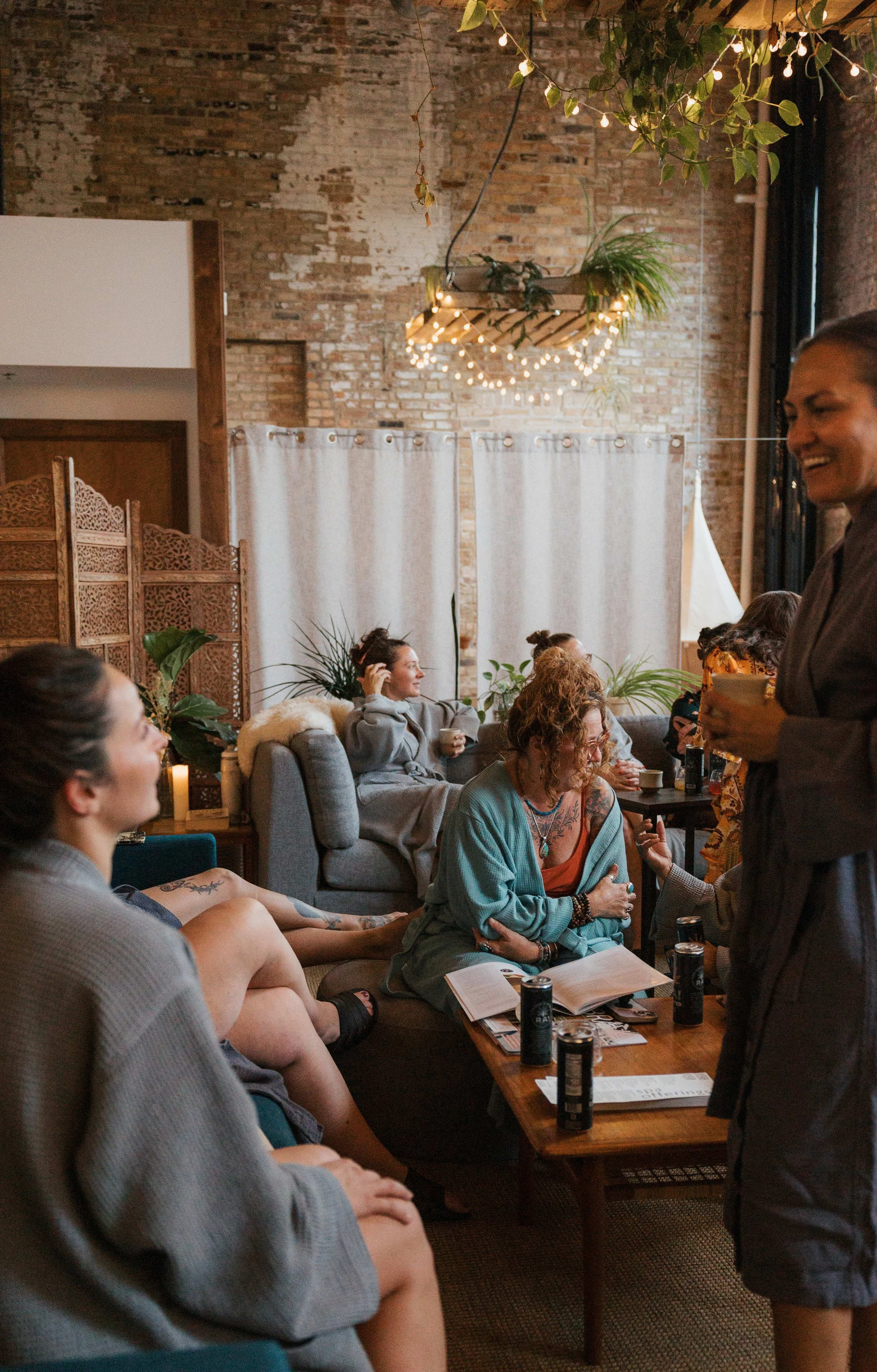Group of women socializing in a cozy indoor space with exposed brick walls, hanging plants, string lights, and a wooden divider. They are engaged in conversation, with some sitting on a sofa, others standing, all smiling.