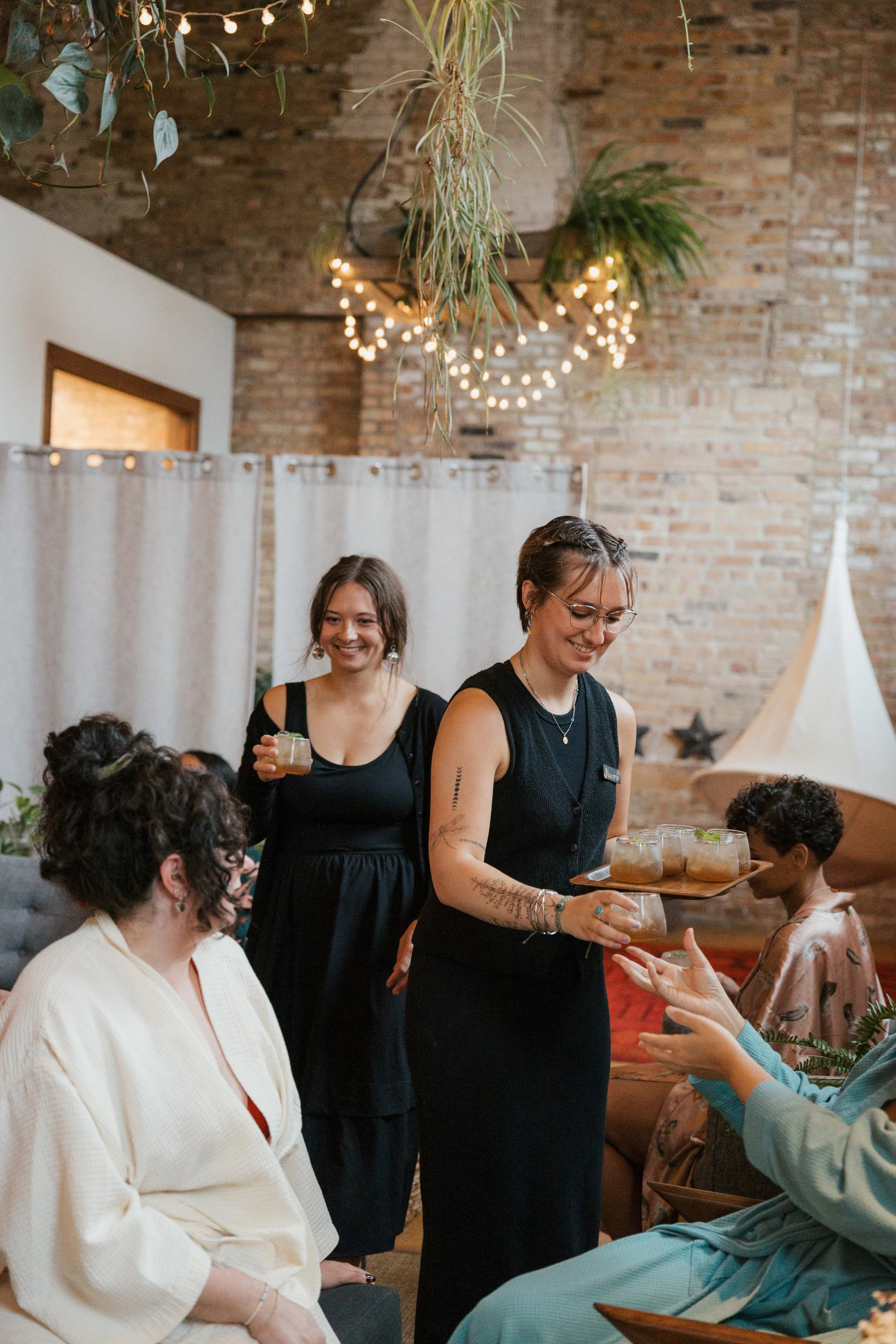 A group of women at a party indoor with a brick wall, string lights, green plants, and a tent in the background. Two women in black is serving drinks, and others are sitting and chatting.