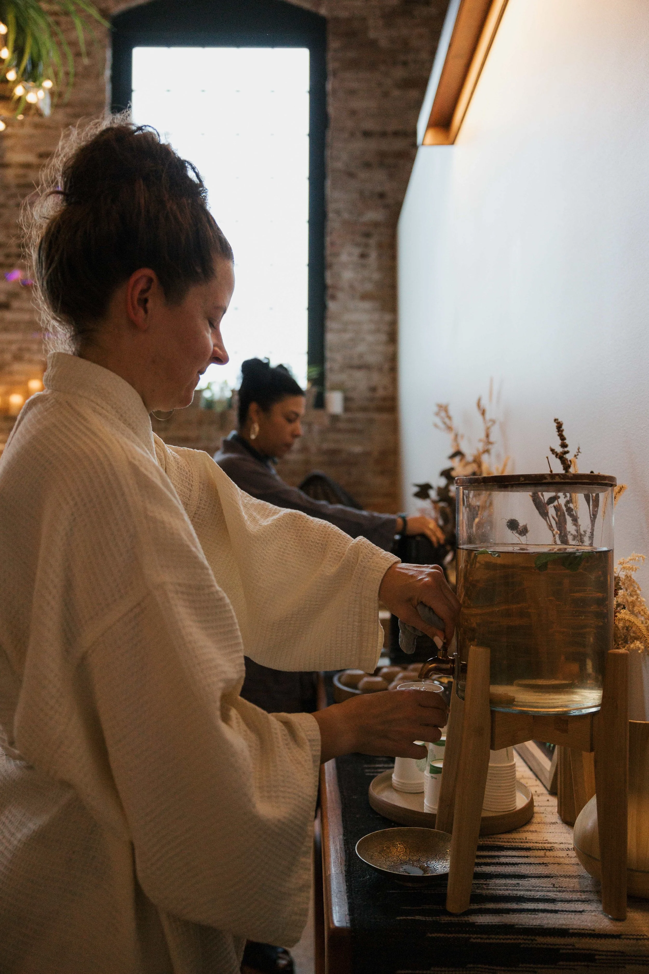 A woman in a beige robe pours a drink from a glass beverage dispenser with water and herbs. In the background, another woman is sitting inside a room with brick walls and large window.