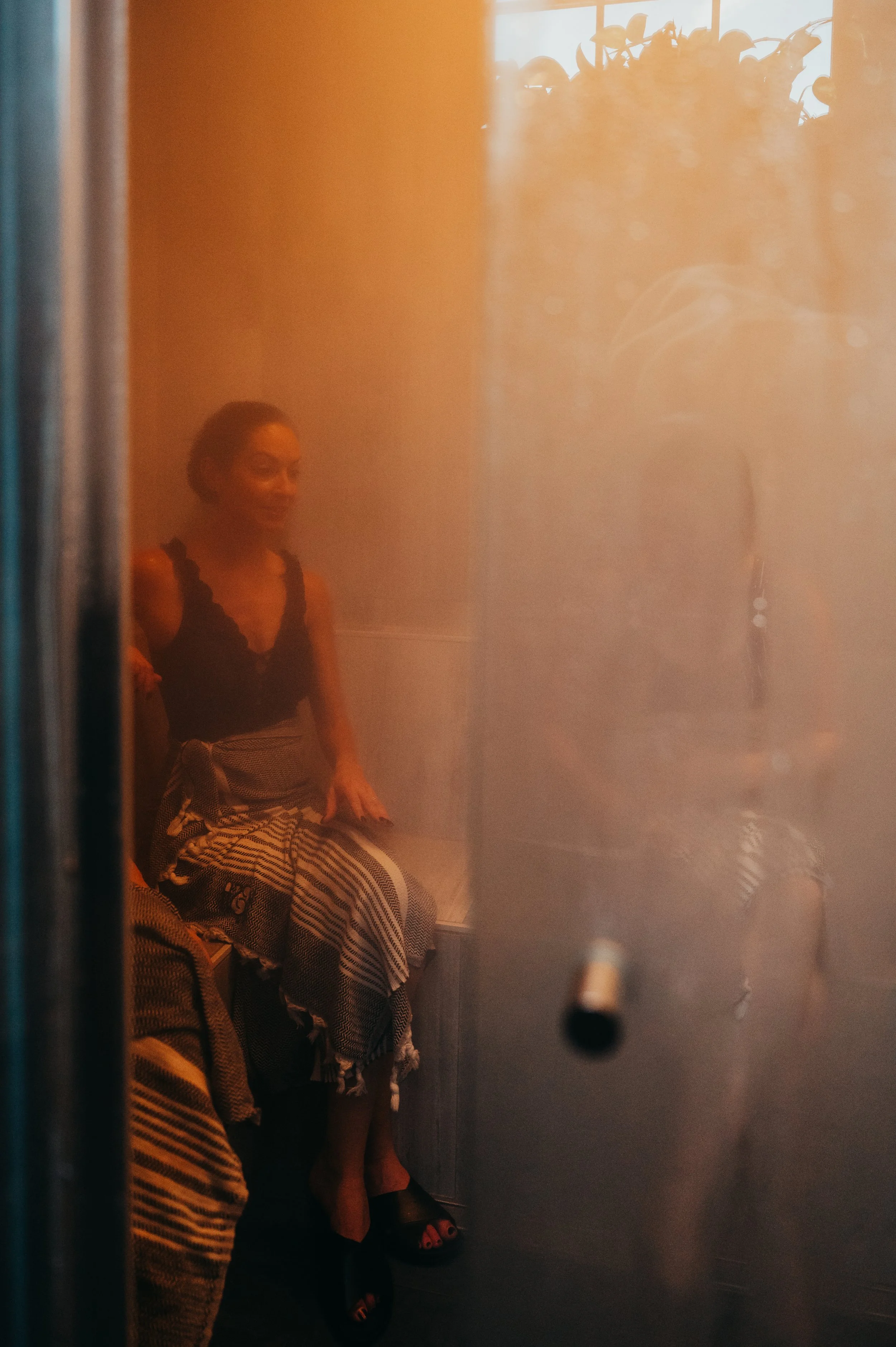 A woman sitting in a dimly lit room, seen through a steam glass door with a reflection of sunlight and plants.