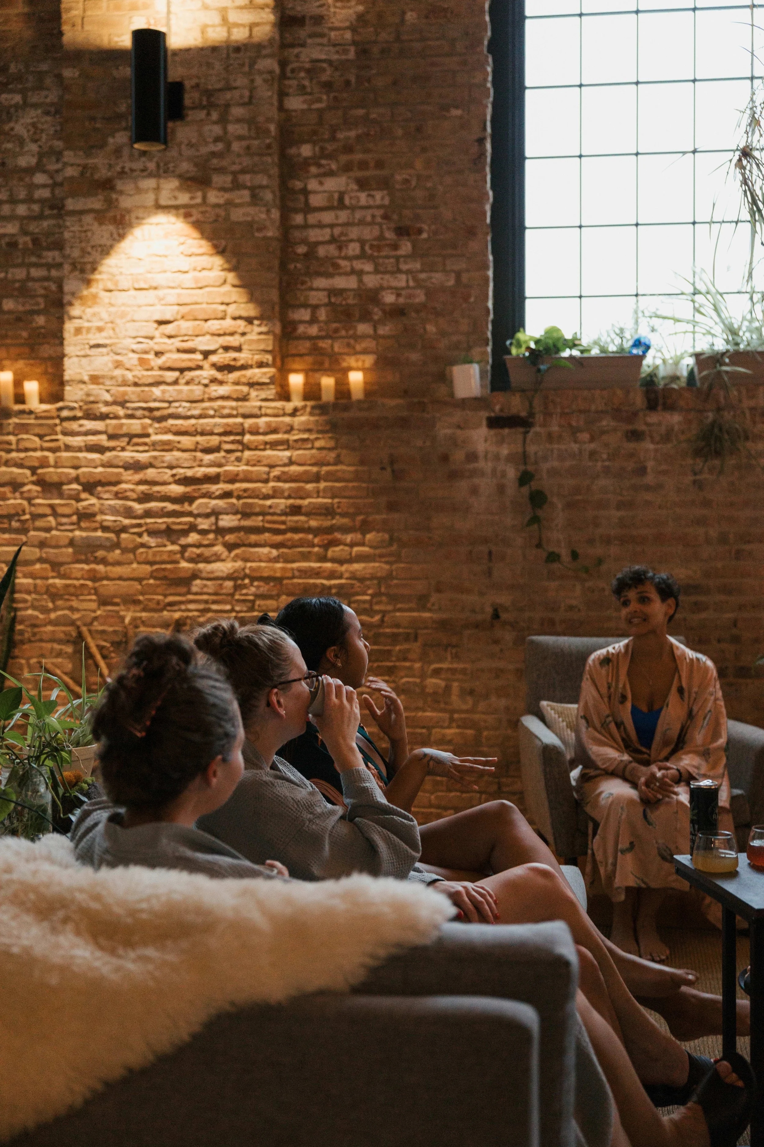 A group of women sitting on couches and chairs having a discussion in a cozy, industrial-style living room with exposed brick walls and large windows.