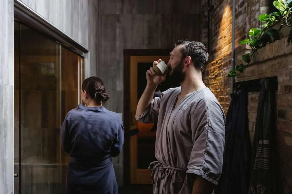 A man with a beard and a gray robe drinks from a white cup in a rustic-style room, while a woman with dark hair tied back and wearing a blue robe stands in the background with her back turned.