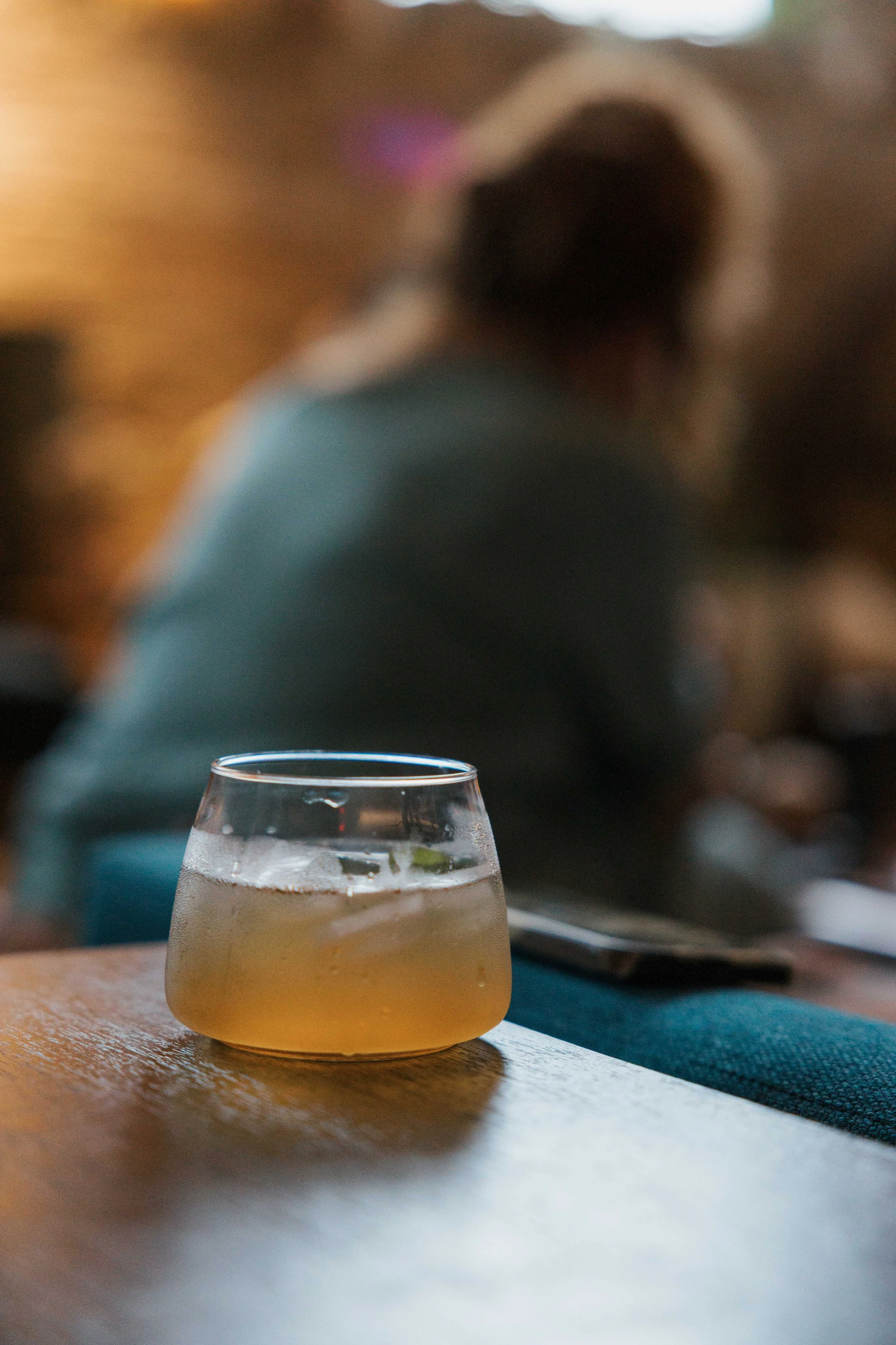 A glass of yellow-colored cocktail with ice cubes on a wooden table in a dimly lit setting, with a blurred person in the background.