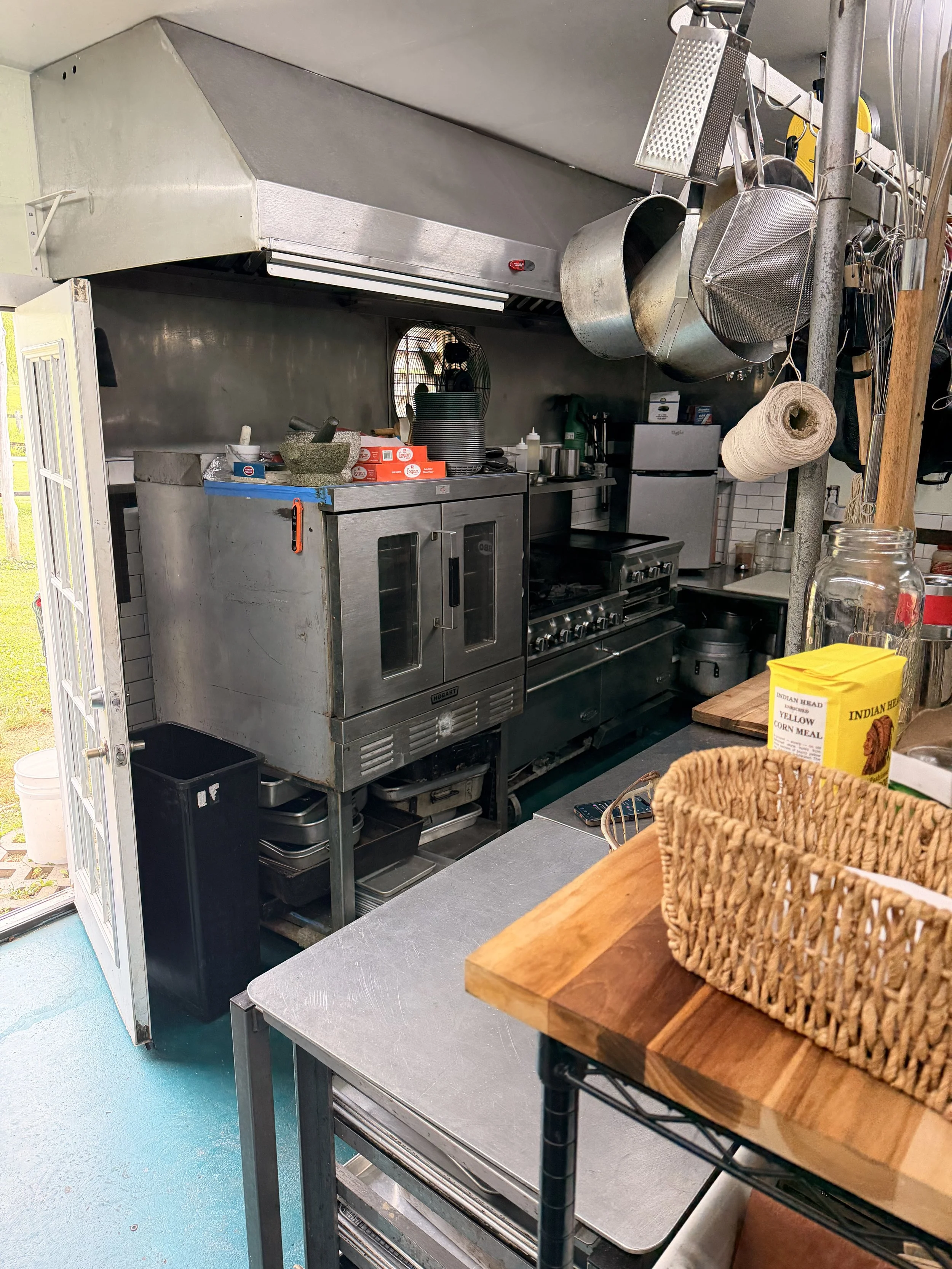 A kitchen with stainless steel appliances and hanging kitchen utensils, including a grater, a colander, and a roll of paper towels, with a woven basket and a yellow bag of Indian Head yellow cornmeal on a wooden counter in the foreground.