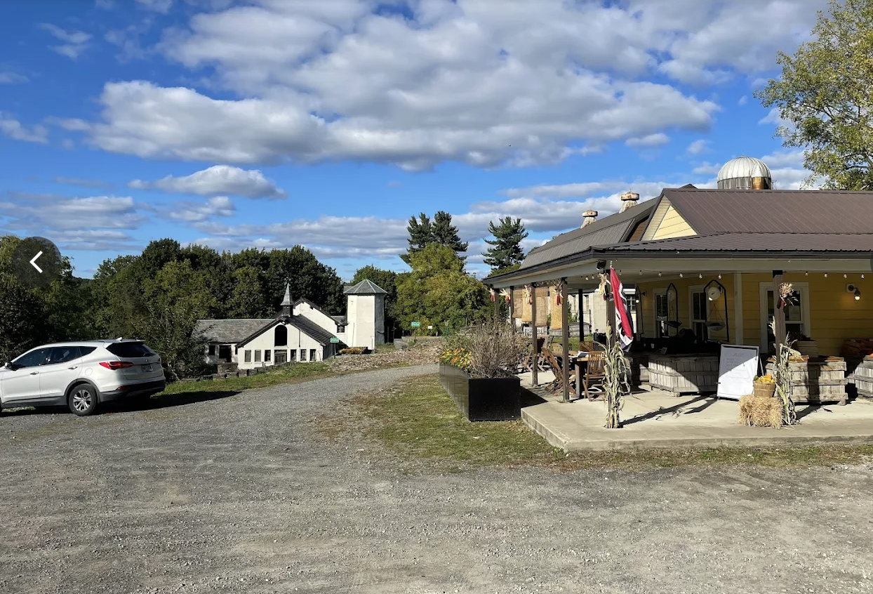 A rural scene with a gravel parking area, a white car, a yellow building with porch seating decorated for fall, a small white chapel or church, and green trees under a partly cloudy sky.