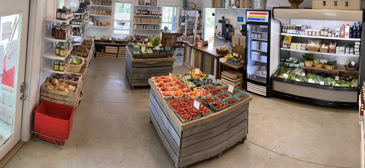 Interior of a grocery store with wooden display tables filled with fresh produce, refrigerators stocked with vegetables and dairy, and shelves with various grocery items.